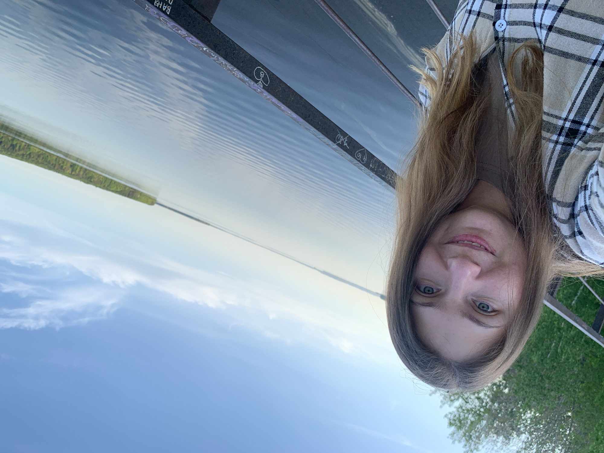 The image shows a selfie of a woman. She has fair skin, light brown hair, and blue eyes. She is wearing a tan and white plaid shirt. The background appears to be a body of water with a thin strip of land on the opposite side. The sky is light blue with some clouds. Part of a metal structure with some graffiti is visible, suggesting a railing or bridge. Greenery is visible to the right of the frame.
Based on the location mentioned (Chaikovsky, Russia), the body of water is most likely the Votkinsk Reservoir. The woman appears to be standing on a viewing platform or bridge overlooking the reservoir.
