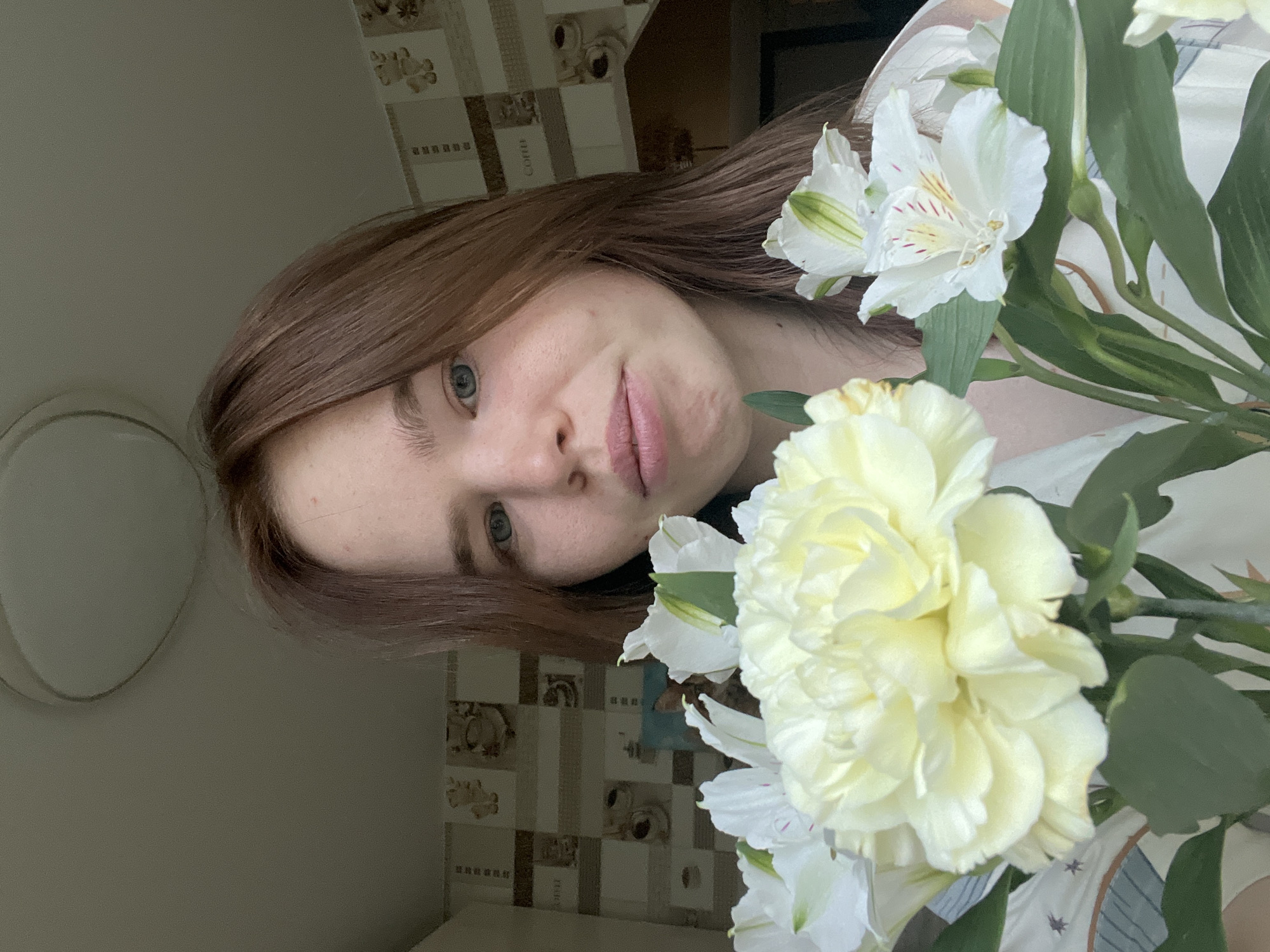 A woman with light brown hair is featured in a close-up shot, holding a bouquet of white and pale yellow flowers. Her face is partially obscured by the blooms. The background reveals a kitchen setting with patterned tiles. The location is Chaikovsky, Russia.