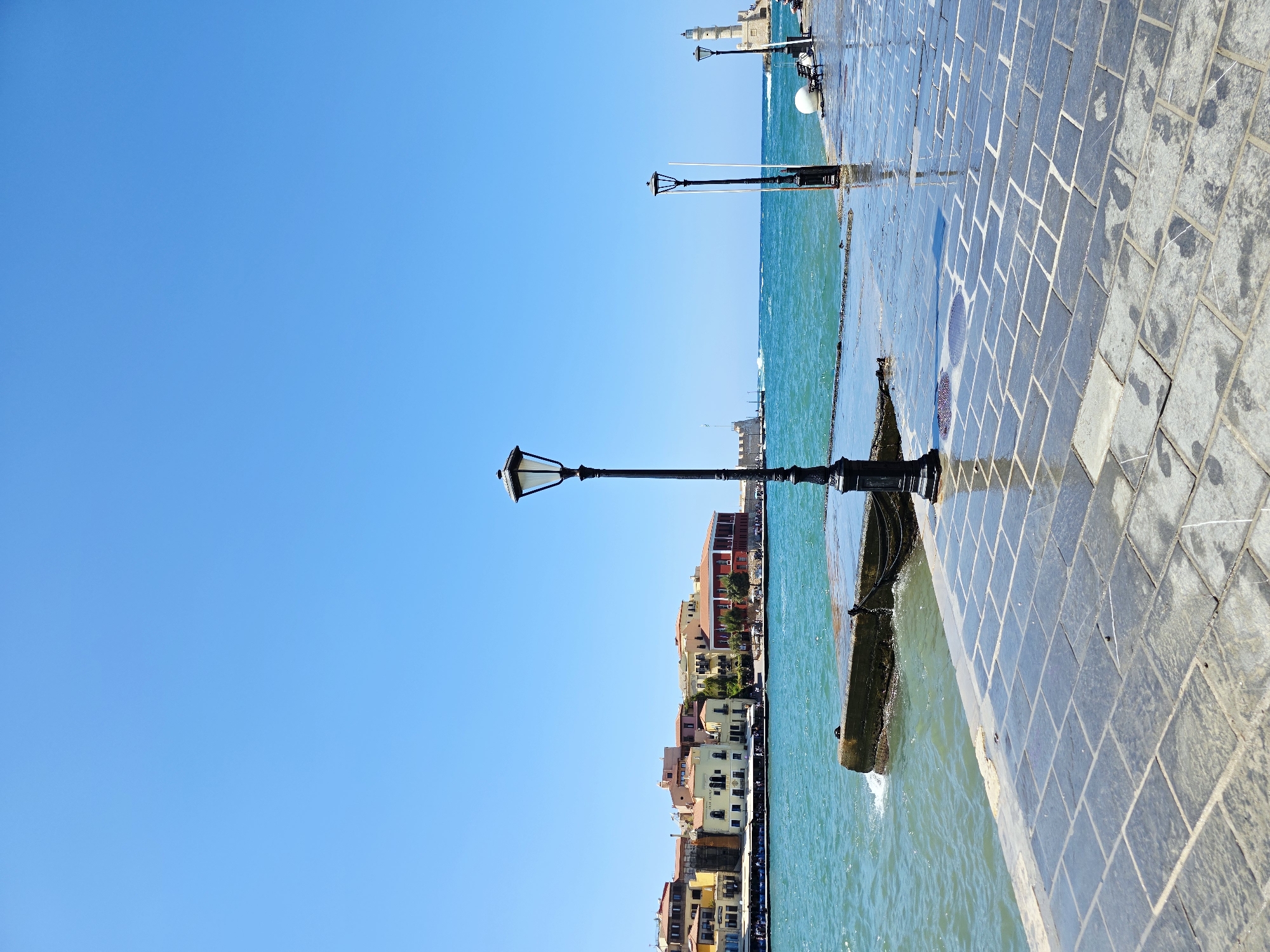 This is a photograph of the Venetian harbor in Chania, Greece, on a clear, sunny day. The foreground is dominated by a wet, stone-paved walkway that reflects the bright blue sky. Several ornate black lampposts line the walkway. To the left, the turquoise water of the harbor laps against a concrete jetty. Across the water, the picturesque old town of Chania is visible, with its colorful buildings, many featuring terracotta roofs. In the distance, a lighthouse stands on a pier, a classic landmark of the harbor. The sky is a vast expanse of uninterrupted blue.