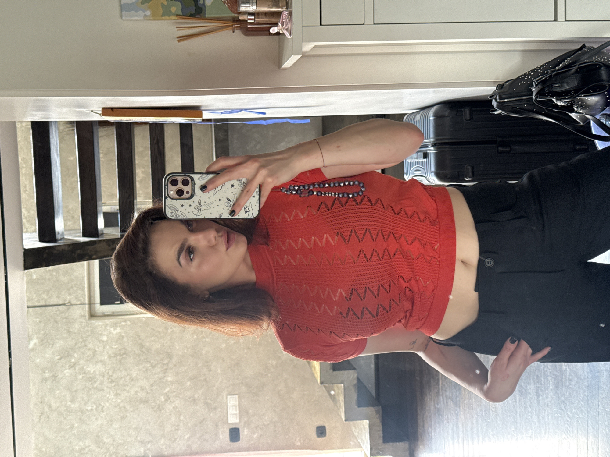 A woman is taking a mirror selfie in what appears to be an apartment. She is wearing a red, short-sleeved, cropped knitted shirt, and black pants. She is holding an iPhone with a patterned case. Behind her in the reflection are stairs, and a piece of luggage. A purse and a cabinet are also visible. There is no specific indication that the location is in Sofia, Bulgaria.
