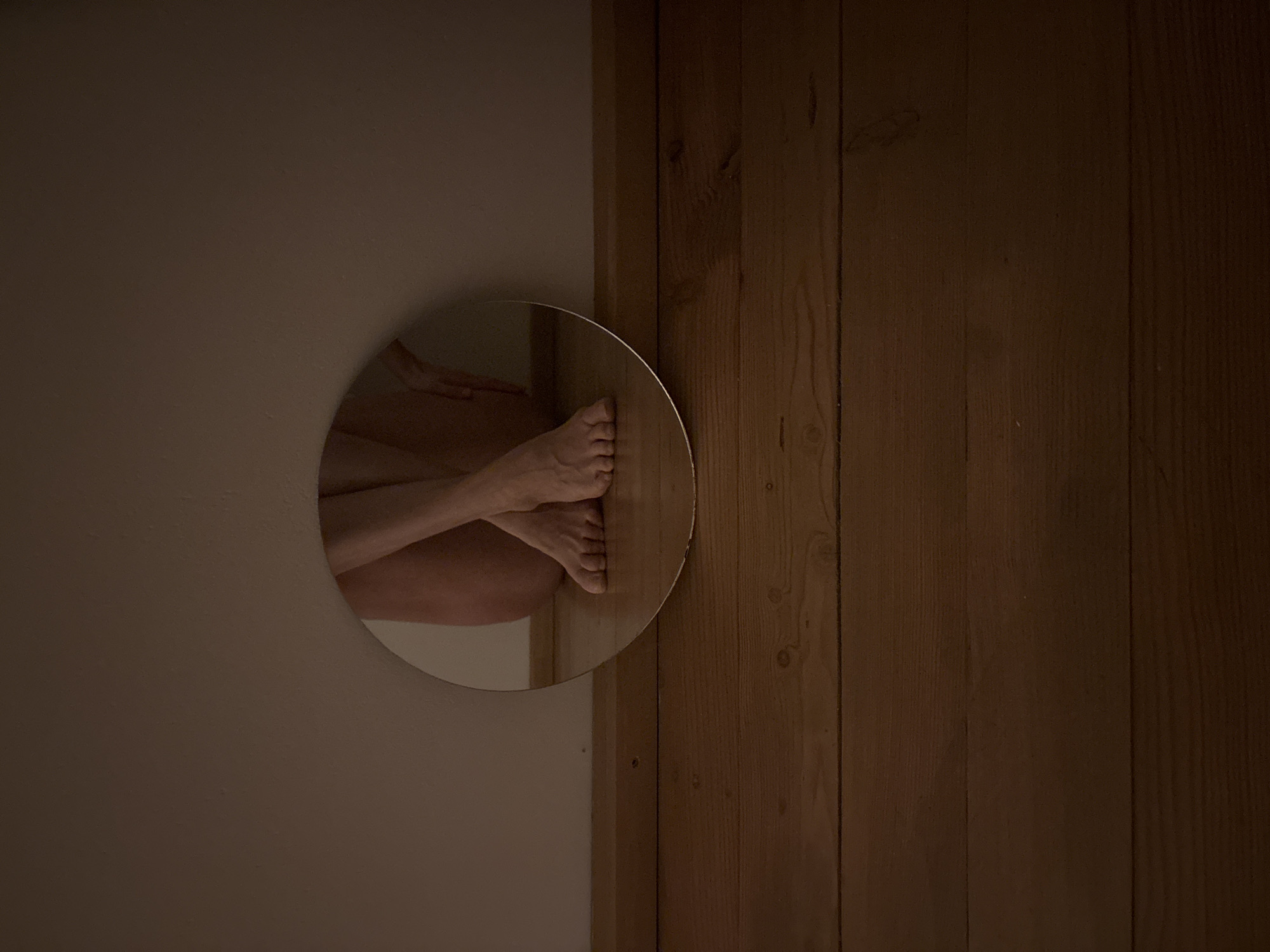 The image shows a round mirror affixed to a beige wall, positioned next to a brown wooden door or wall paneling. Reflected in the mirror are human legs and feet. The setting is ambiguous; there is no readily identifiable information within the image to pinpoint the location as Chaikovsky, Russia.