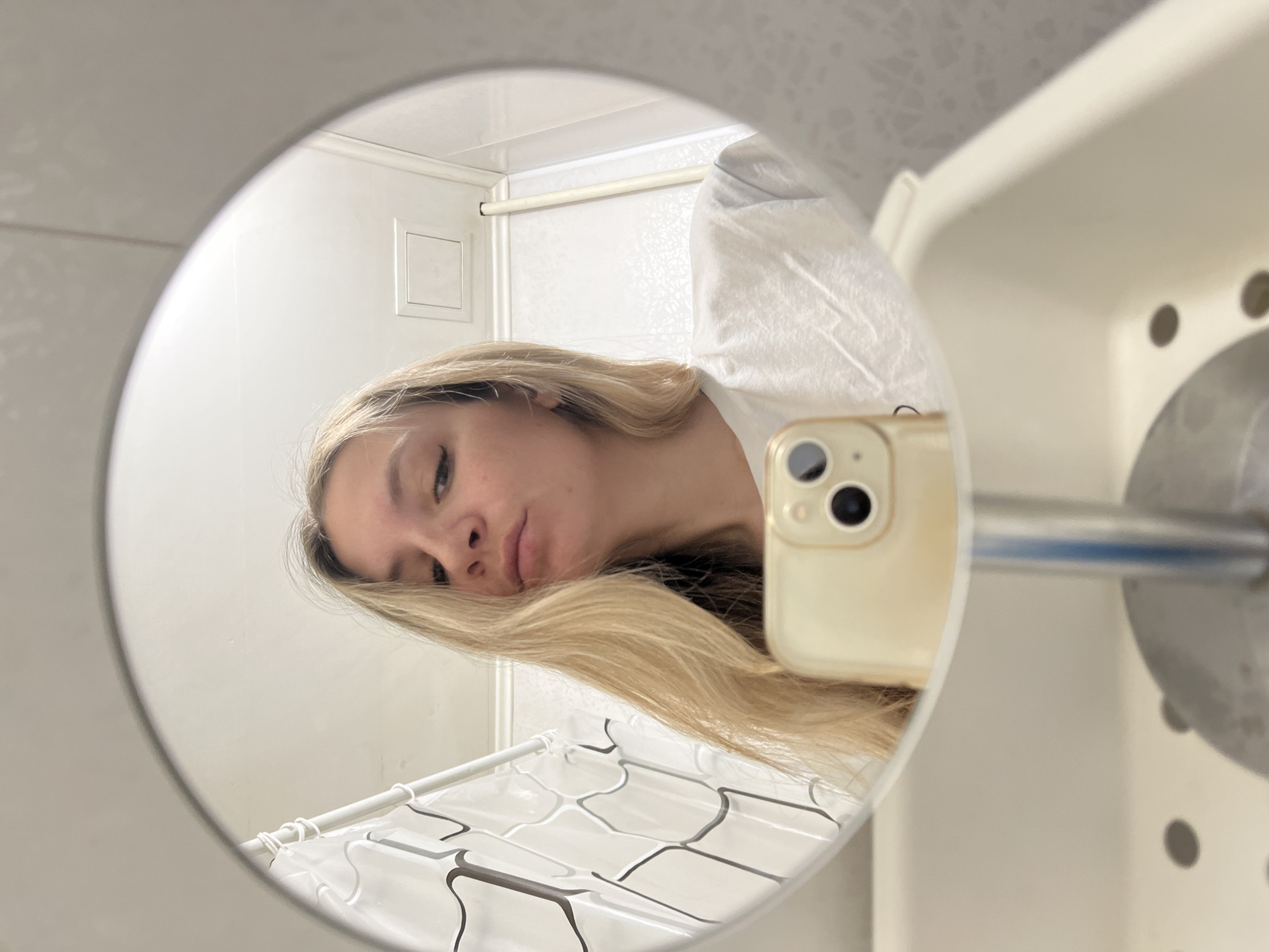 An indoor, close-up image captures a woman taking a mirror selfie in what appears to be a bathroom. The central subject is the reflection of a woman with long, straight blonde hair, fair skin, and a neutral or slightly pensive expression, looking directly into the camera. She is wearing a white t-shirt. Her right cheek and ear are partially obscured by a light-colored smartphone (likely an iPhone) she is holding, which has a distinct camera lens configuration.
The reflection in the circular mirror shows parts of a bathroom: a white shower curtain with a subtle geometric pattern, hanging from a white shower rod at the bottom, and a white tiled wall with a small square access panel in the background. Around the mirror's edge, outside the reflection, visible elements include a white wall or ceiling, and to the right, a portion of a white plastic storage unit with circular holes, from which a metallic rod extends towards the mirror. The overall lighting is bright, suggesting indoor illumination. There is no visible text in the image. The time of day and weather are not discernible.
