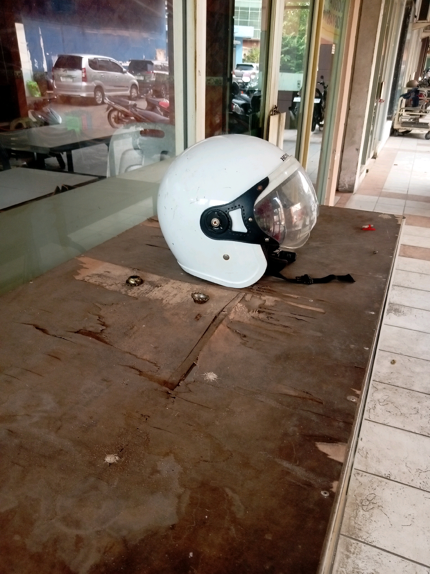 A white motorcycle helmet with a clear visor sits on a weathered, dark brown wooden table. The helmet has a black chin strap and a black trim around the visor. The table surface is damaged and shows signs of wear, with visible wood grain and some debris. In the background, reflections in a large glass window show a silver van, several motorcycles, and storefronts. To the right of the table, a tiled walkway and the exterior of a building are visible. The scene appears to be outdoors, possibly in a commercial area in Batam, Indonesia.