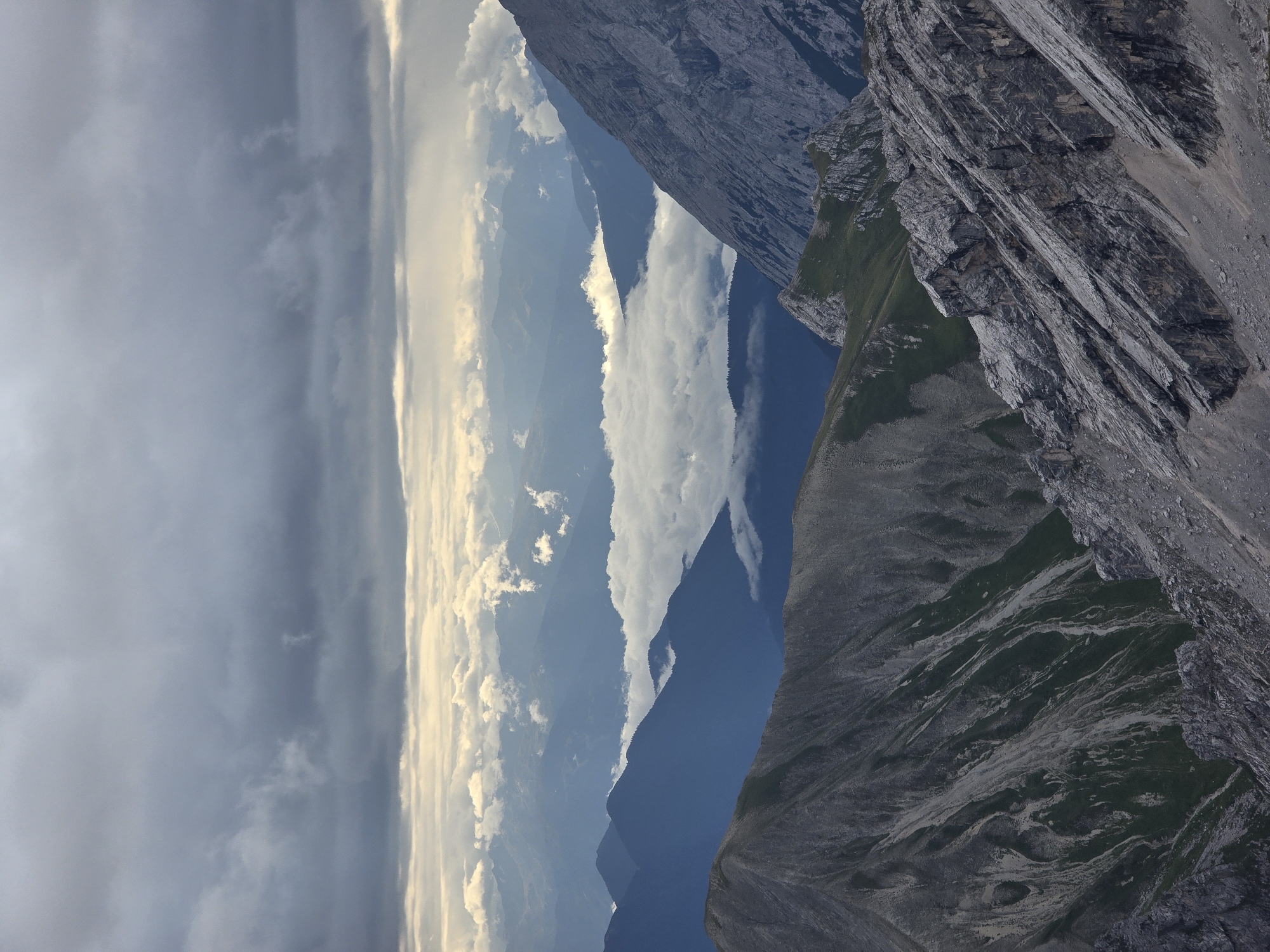 The image is a vertical, medium-distance, outdoor photograph of a mountainous landscape under a cloudy sky. The foreground features rugged, rocky slopes with patches of green vegetation. In the mid-ground, layers of blue-hued mountains recede into the distance, interspersed with a band of white clouds that appear to be a cloud inversion. The background consists of more distant mountain ranges shrouded in haze. The sky is dominated by heavy, dark gray clouds, with some lighter patches suggesting breaks in the cloud cover. The scene is devoid of people or any man-made structures, presenting a raw and natural environment. The lighting is subdued due to the overcast conditions.