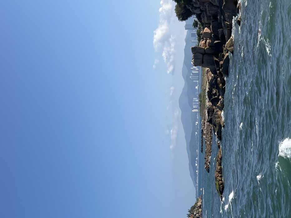 The image displays a coastal scene in Nha Trang, Vietnam. A vast, clear light blue sky occupies the upper two-thirds of the frame, with minimal scattered white cumulus clouds visible above the distant horizon. The lower third features dark blue-green ocean water with visible white-capped waves. Numerous rugged, dark brown and black rock formations protrude from the water and line the immediate shore. On the far left, a small rocky island or headland, partially covered with green vegetation and featuring a small, light-colored structure at its peak, is visible. Two small, white-colored boats are present in the water near this headland. In the mid-right, a prominent cluster of large, dark rocks is stacked, with smaller indistinct figures, likely people, walking or standing on a sandy-grassy area behind them. White specks, consistent with birds, rest atop the largest rock formation. The background presents a long, hazy range of dark blue-grey mountains. In front of the mountains, a distinct skyline of numerous modern high-rise buildings indicates an urban coastal environment. Sparse green vegetation is visible on the rocky terrain and distant land.