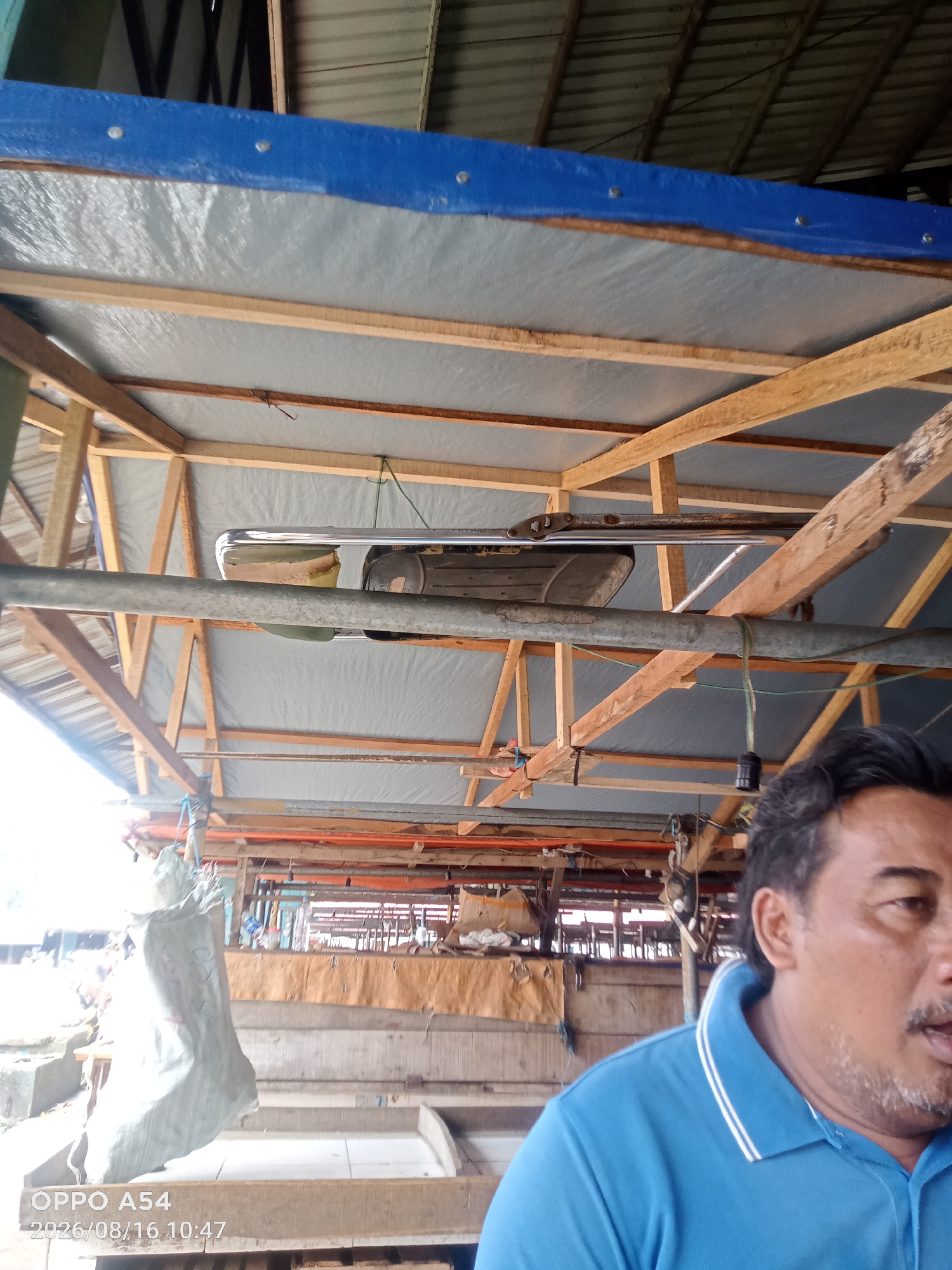 The image shows a close-up of a man wearing a blue polo shirt with white trim. He has dark hair and a mustache and is looking to the right. Behind him, there is a wooden structure with a blue tarp and a silver object, possibly a piece of machinery, suspended from it. The background is blurred but appears to be an outdoor market or workshop setting, with other structures and possibly goods visible. The timestamp in the lower left corner indicates the photo was taken on 2026/08/16 at 10:47. This scene is consistent with an outdoor stall or workshop environment, commonly found in markets throughout Jambi, Indonesia.
