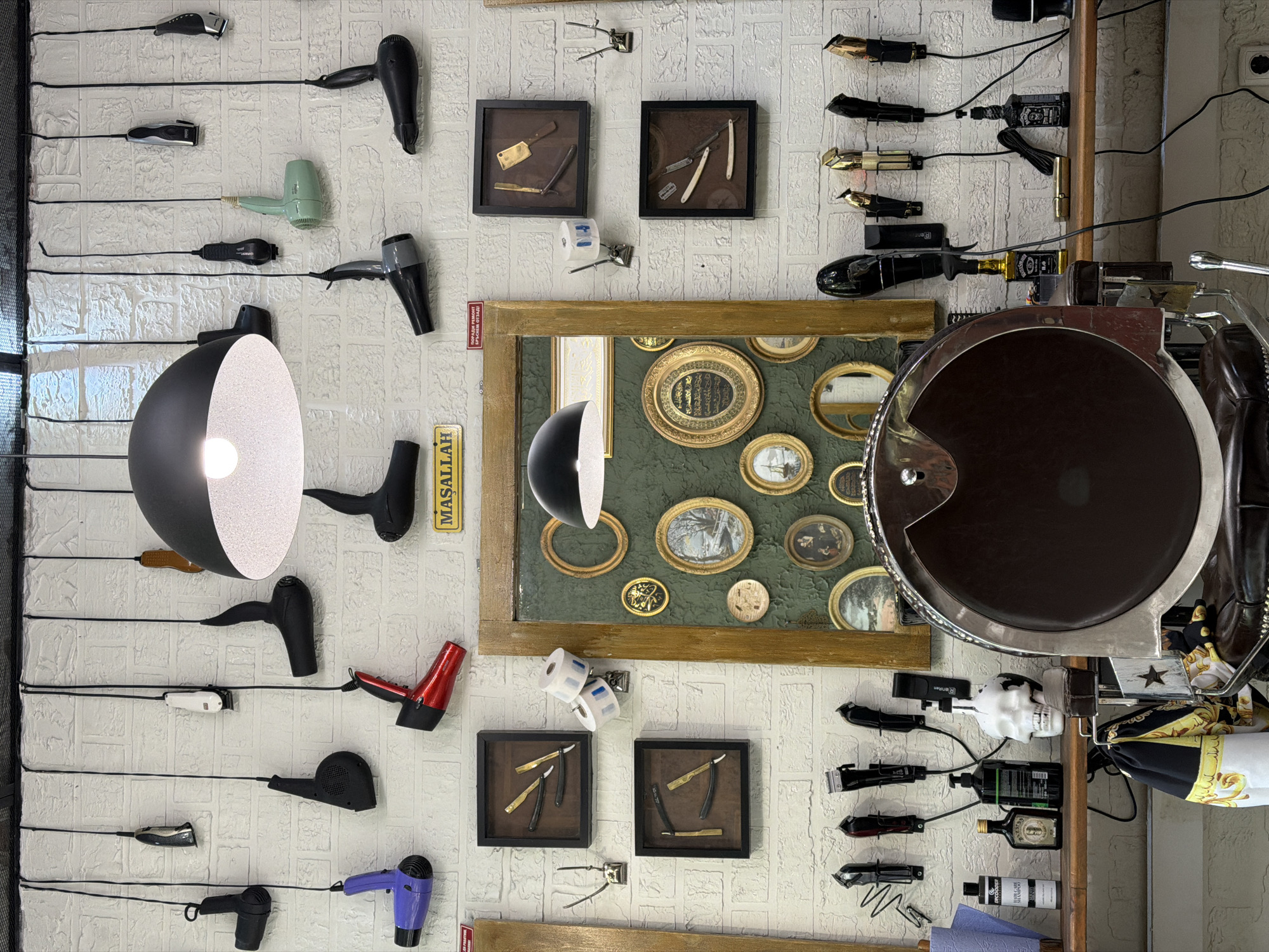 The image shows a wall in what appears to be a barbershop or hair salon. The wall is made of white textured bricks and is adorned with various objects related to the barbering trade.
There are several hair dryers and hair clippers mounted on the wall, arranged in rows and columns. Some of the hair dryers are vintage models, while others are more modern. The hair clippers are also a mix of old and new styles.
In addition to the hair dryers and clippers, there are also several framed displays on the wall. These displays contain antique straight razors and other barbering tools. There is also a large framed collection of small, oval portraits of people.
Other items on the wall include a lamp, a sign written in Turkish which states "MASALLAH" (which is an Arabic phrase used to express appreciation, joy, praise, or thankfulness for an event or person that was just mentioned, or a general expression of good will), a skull, and several bottles of hair products. A barber chair with equipment is partially visible on the right side of the image.
While I cannot confirm the exact location, it's mentioned that the city is Sofia, Bulgaria. Without additional specific details visible in the image, I cannot confirm the city.