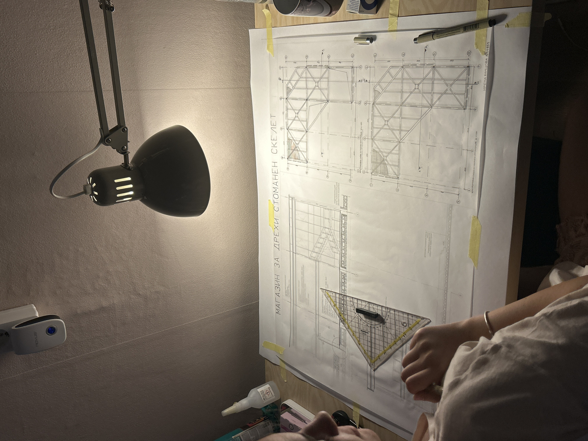The image shows a close-up view of a person's hands and part of their torso as they work on architectural blueprints spread across a wooden desk. The blueprints, detailing a "MAGAZINE FOR CLOTHING STEEL FRAME," are secured with yellow tape. A drafting triangle and a pen are positioned on the plans. A desk lamp with a green metal shade illuminates the workspace. In the background, a white object with a blue light, possibly a motion detector or a sensor, is attached to a textured, sloping ceiling. The overall environment appears to be a home office or studio, likely within the city of Sofia, Bulgaria, given the Bulgarian text on the blueprints and the common use of such equipment in architectural practices in the region.