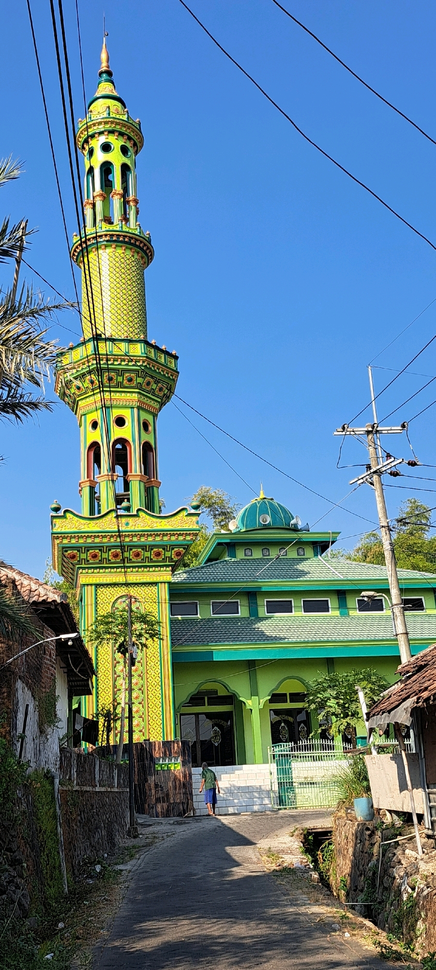 A vibrant, green mosque with a tall, elaborately decorated yellow and green minaret stands under a clear blue sky in Candi Wates, Indonesia. The minaret is cylindrical with multiple levels of intricate patterns, balconies, and decorative arches. The main mosque structure is also green, featuring a green tiled roof, a smaller green dome, and numerous rectangular windows. A man wearing a green shirt and blue shorts walks away from the mosque up a paved path. The path is bordered by stone walls and vegetation, with power lines visible crisscrossing the sky. Several houses with rustic tile roofs are visible on either side of the path, partially obscuring the mosque and minaret from view. The scene depicts a sunlit day in a semi-urban or village setting.