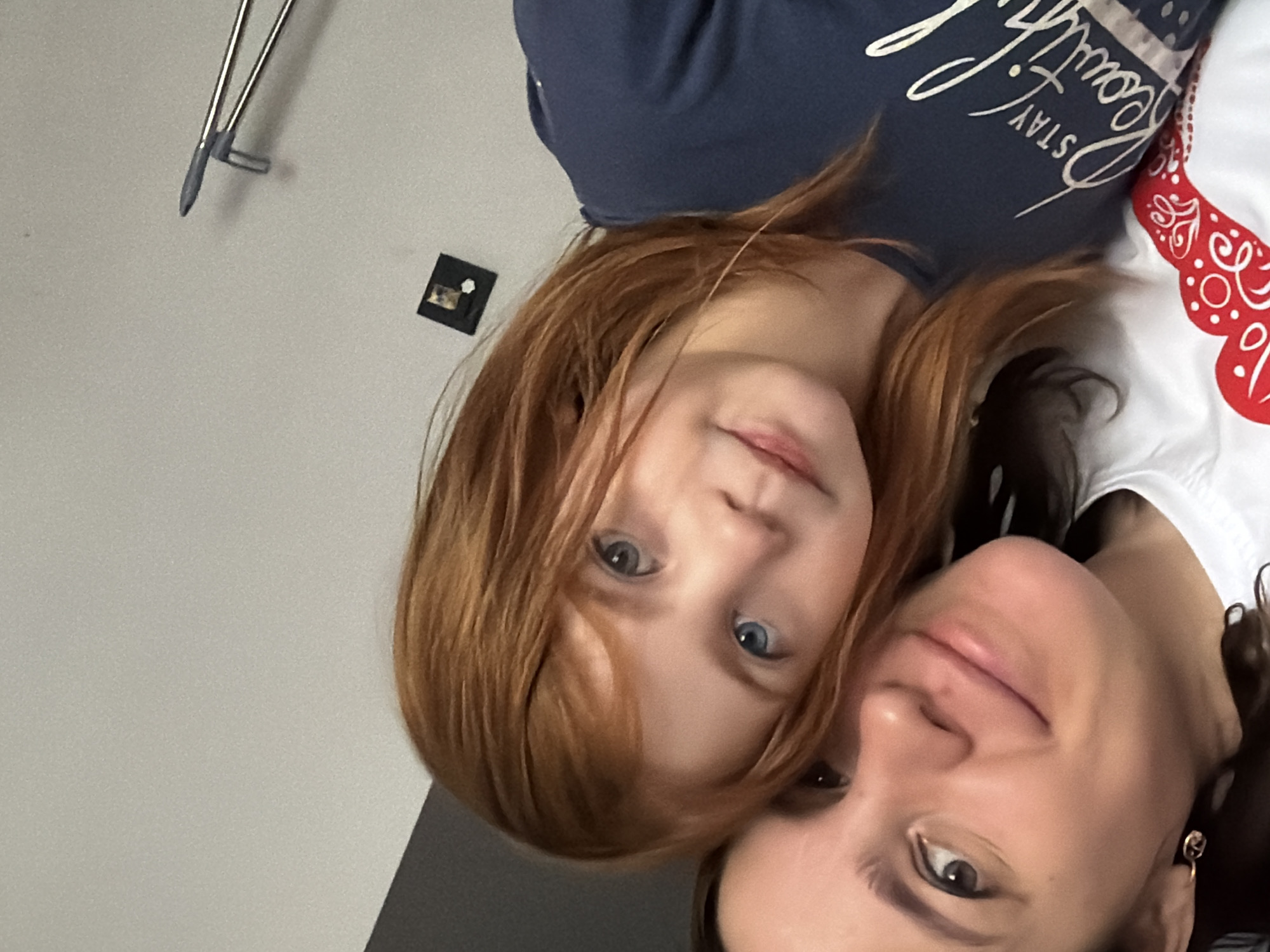 In a self-taken photograph, a woman and a young girl with red hair are close together, facing the camera. The woman, on the left, is smiling, her left ear adorned with a small, gold hoop earring. She is wearing a white t-shirt with a red, decorative pattern. The girl, positioned to the right of the woman, has striking blue eyes and her red hair frames her face. She is wearing a dark blue t-shirt with white lettering that reads "STAY Beautiful". The background consists of a plain, light-colored wall with a small, dark framed object hanging on it, and further to the right, the angled tip of a metallic object, possibly tweezers or a similar tool. There are no discernible landmarks or contextual clues within the image to suggest a specific location in the city of Perm, Russia, or any other city. The lighting appears to be ambient indoor lighting.