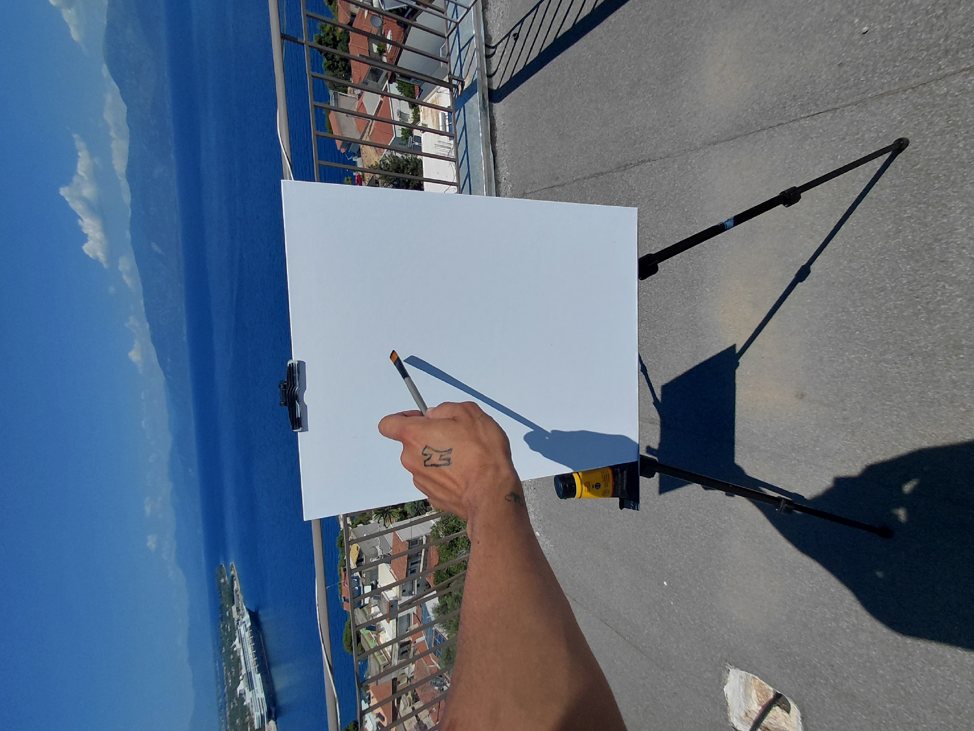 The image depicts an outdoor painting setup with a person's arm and hand holding a paintbrush poised over a blank canvas on an easel. The scene is set on a rooftop or elevated balcony overlooking a body of water and a coastline. In the distance, mountains form a backdrop. A large cruise ship is visible anchored in the blue water near a coastal town with red-tiled roofs. A small yellow bottle of art supply is placed on the easel. The lighting suggests a sunny day, casting shadows on the textured ground. The location is within the Municipal Unit of Aegio, Greece.