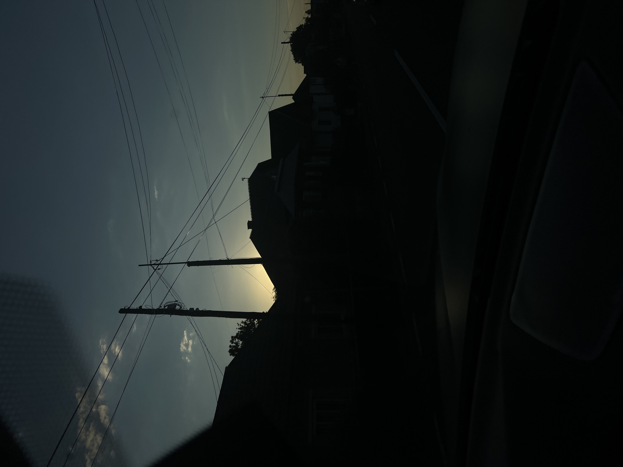 The image depicts a street scene at dusk in Rona de Sus, Romania. Silhouetted against a darkening sky, several houses line the street, their architectural details obscured by the low light. Overhead, a network of power lines stretches across the frame, supported by utility poles. The sun is low on the horizon, casting a warm glow behind a cluster of trees and the rooftops of the houses, creating a dramatic silhouette. The foreground is dominated by the dark interior of what appears to be a vehicle, with a glimpse of the dashboard and windshield visible. The overall mood is quiet and somewhat somber, characteristic of twilight in a residential area.