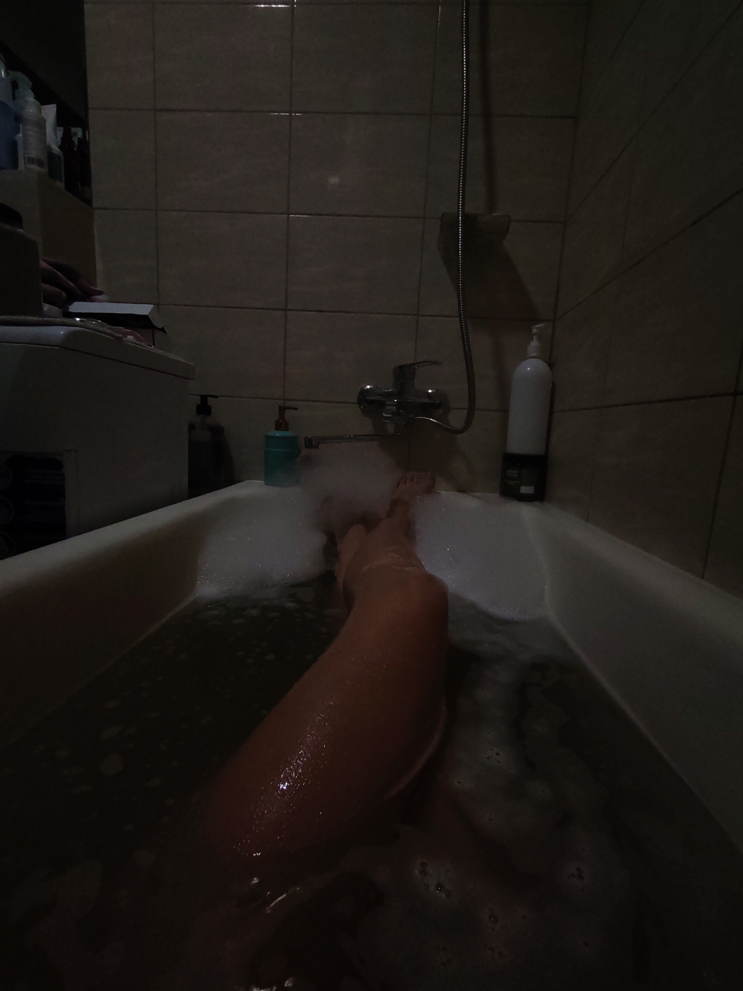 In a dimly lit bathroom, likely located in Izhevsk, Russia, a person's leg is submerged in a bathtub filled with foamy water. The bathtub is made of white material and is surrounded by tiled walls. On the side of the tub, near the faucet, are several bottles of toiletries, including a tall white bottle with a pump dispenser. A shower head is visible mounted on the wall. The lighting in the room is low, casting shadows and highlighting the bubbles in the water. The scene appears to be a private moment of relaxation.
