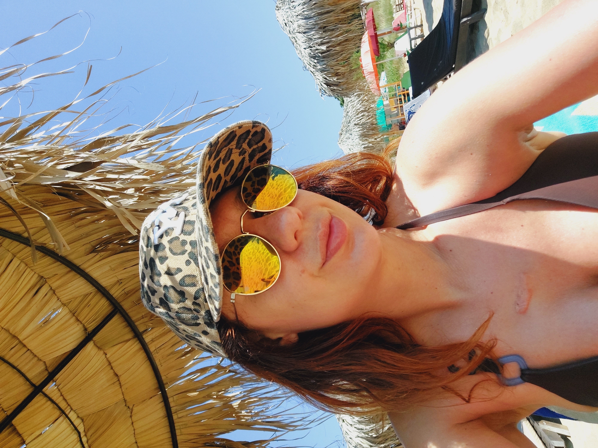 A woman with reddish-brown hair and a leopard-print baseball cap is taking a selfie on a beach. She is wearing sunglasses with yellow, reflective lenses and a dark bikini top. Her left arm and chest are visible, showing a scar on her chest. She is sitting under a thatched umbrella, with more thatched umbrellas and beach chairs visible in the background. The sky is blue, and the scene appears to be in a coastal resort area. The location is the beach of Sozopol, Bulgaria.