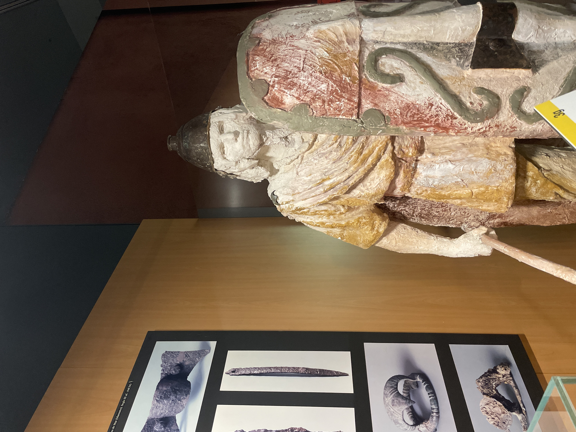 A tilted, overhead shot captures a museum display. The primary focus is a detailed, painted sculpture depicting a human figure, possibly a warrior or noble, lying on a bed or bier. The sculpture is painted in earthy tones of white, brown, and gold, with hints of red, green, and silver on the head and details on the top. The figure appears weathered and aged.
Beneath the sculpture, on a light brown surface, is a display panel featuring photographs of what appear to be archaeological findings. The photographs showcase artifacts, including what seems to be a sword, jewelry, and fragments of other metallic objects.
A small yellow label is visible near the top of the sculpture. Text visible on one of the labels of one of the archaeological findings, “ra de Mar (Maresme). Segle III aC. (MCM).”
Based on this information, it is an exhibition of artifacts of the city of Barcelona, Spain.