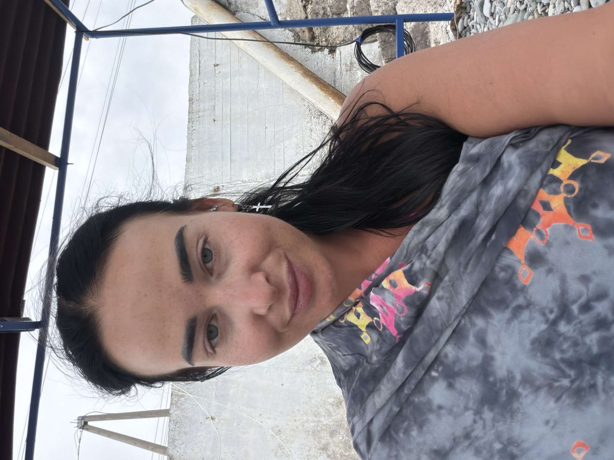 A woman with long dark hair and light eyes is pictured from the chest up, looking directly at the camera with a slight smile. She is wearing a grey tie-dye wrap with colorful geometric patterns. A small silver cross earring is visible in her left ear. The background appears to be an outdoor setting with a concrete wall, a metal structure, and a cloudy sky. This image was taken in Sochi, Russia, at the Black Sea coast, potentially near a beach or a coastal promenade. The woman's expression is relaxed and natural, suggesting a casual moment captured on camera.