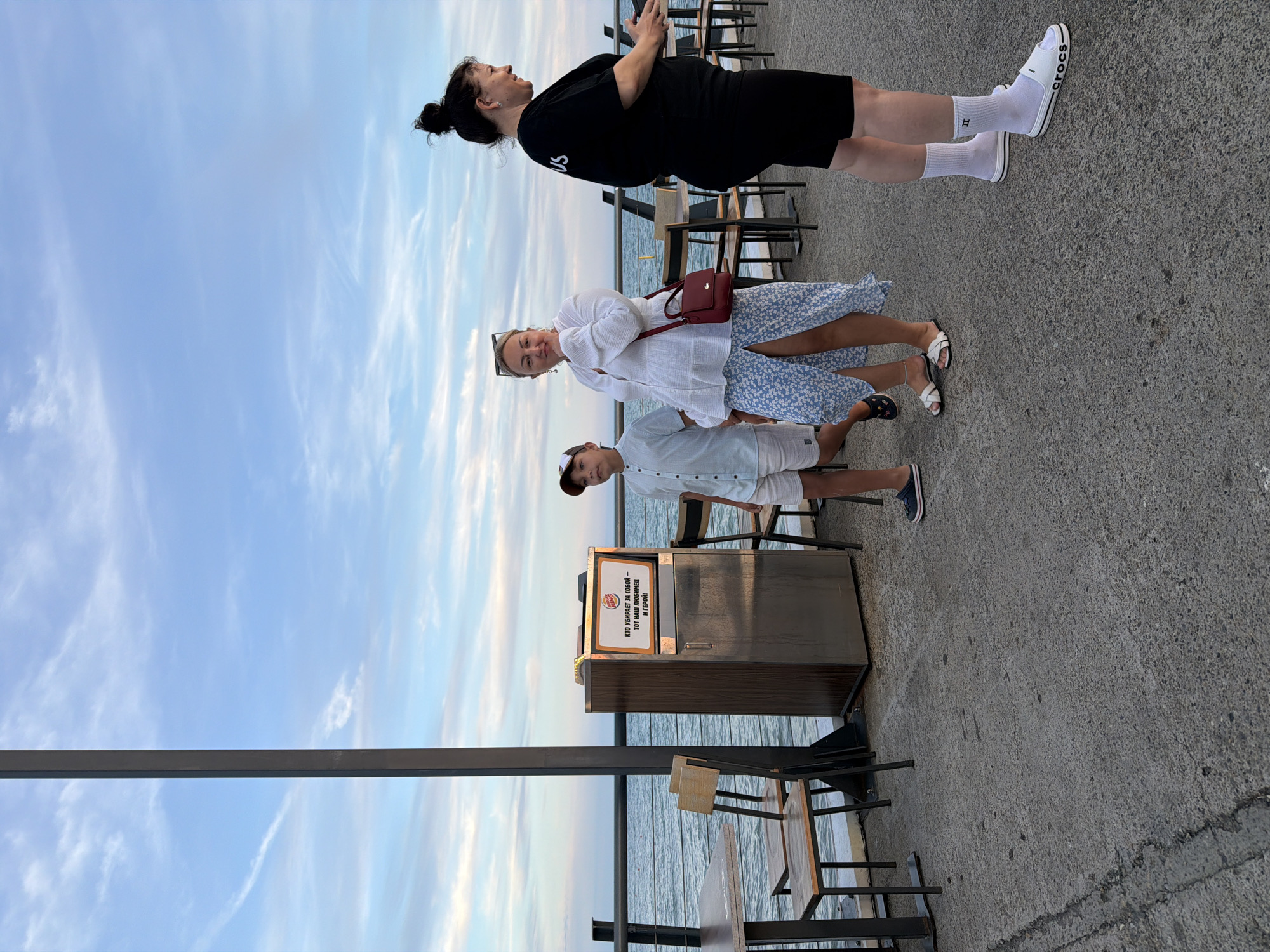 The image depicts a group of people on a promenade next to the sea. A young boy in a light blue shirt and white shorts stands to the left, looking towards the viewer. Next to him, a woman in a white blouse and a blue floral skirt is looking over her shoulder with a slight smile, holding a red purse. To the right, another woman in a black dress and white socks with black slides stands facing away from the camera, her head turned to the side. In the background, tables and chairs are set up, and a trash receptacle with a Burger King sign is visible. The scene appears to be set in Sochi, Russia, given the architectural style and the presence of the sea. The sky is a pale blue with scattered clouds, suggesting it is either early morning or late afternoon. The overall atmosphere is casual and relaxed, with people enjoying a seaside outing.