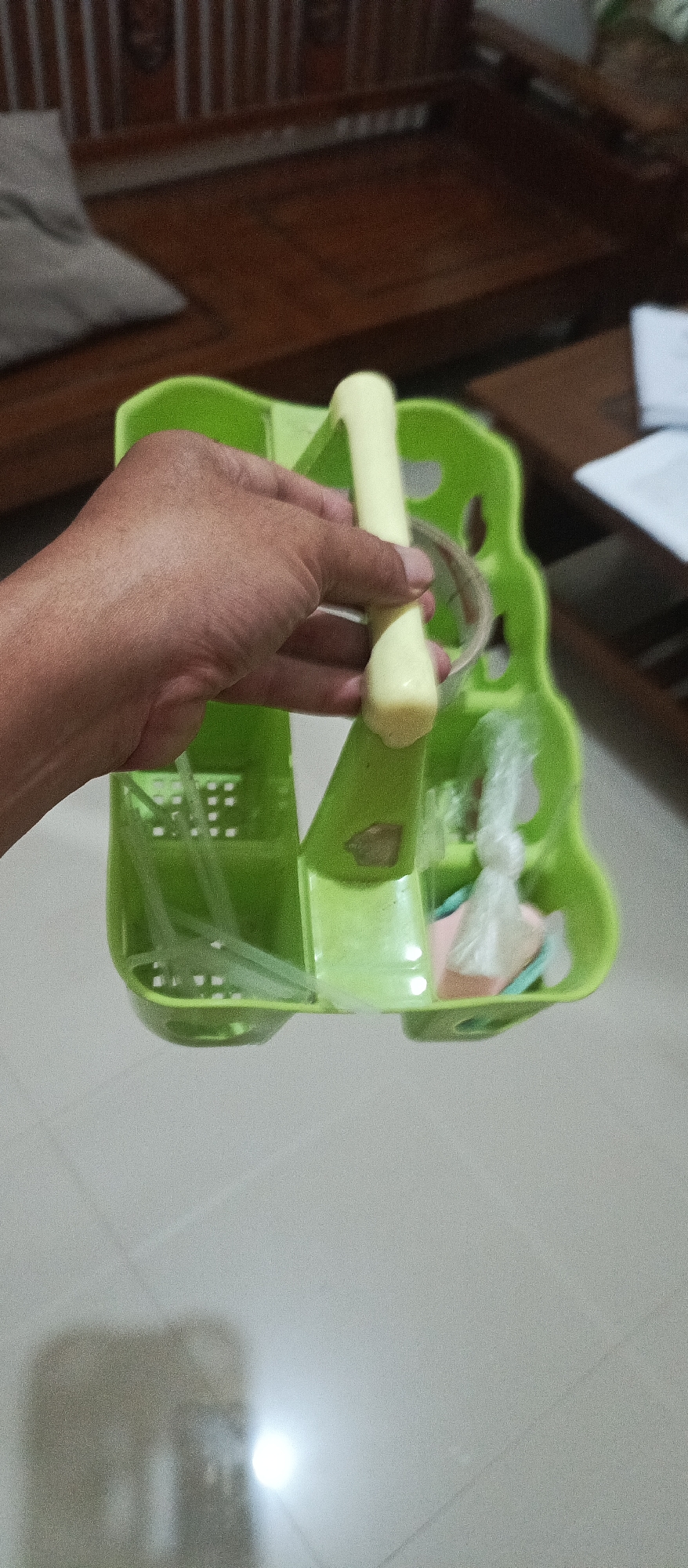 An overhead shot shows a person's left hand holding the light yellow handle of a green plastic caddy. The caddy has a wavy top edge and multiple compartments. Inside one compartment, thin, translucent plastic straws are visible. Another compartment contains a white plastic bag tied with a string, and under it are small, colorful items, possibly paper or fabric. The caddy is positioned in front of a blurry background of what appears to be wooden furniture and a tiled floor, suggesting an indoor setting in a home or establishment. The lighting suggests the image was taken indoors with artificial light. This scene is located in Arjasa, Indonesia.