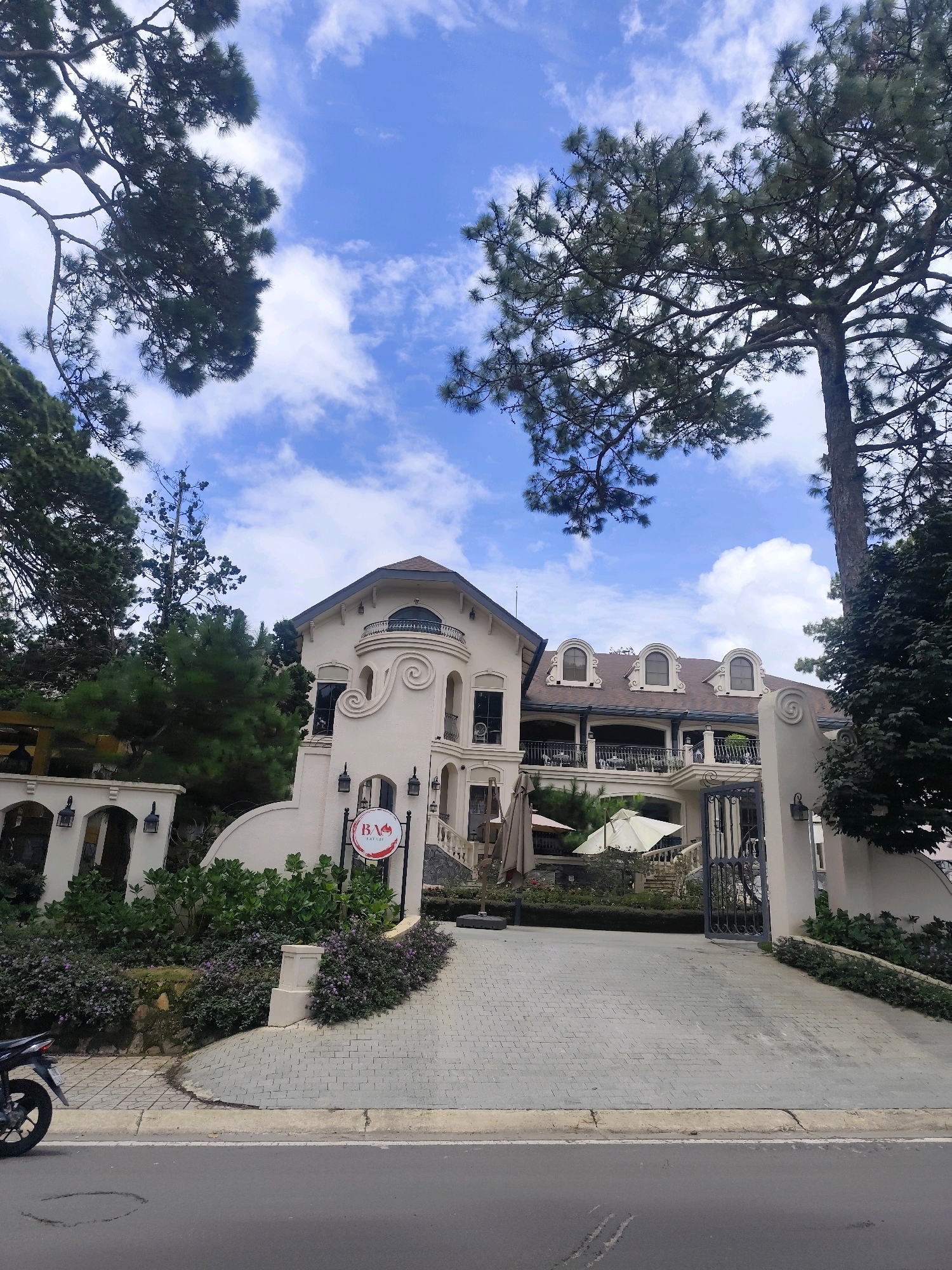 The image depicts a large, cream-colored villa situated in Lâm Viên - Đà Lạt, Vietnam. The villa features ornate architectural details, including curved facades, arched windows, and decorative moldings. The roof is dark brown with dormer windows. In front of the villa, there is a paved driveway leading to a wrought-iron gate. Lush greenery, including trees and flowering bushes, surrounds the property. The sky above is blue with scattered white clouds. In the foreground, a portion of a paved road is visible, with a motorcycle parked on the left side. A circular sign with red lettering is attached to a post near the entrance of the villa. The overall impression is one of a grand and well-maintained property, characteristic of the highland architecture found in Đà Lạt.