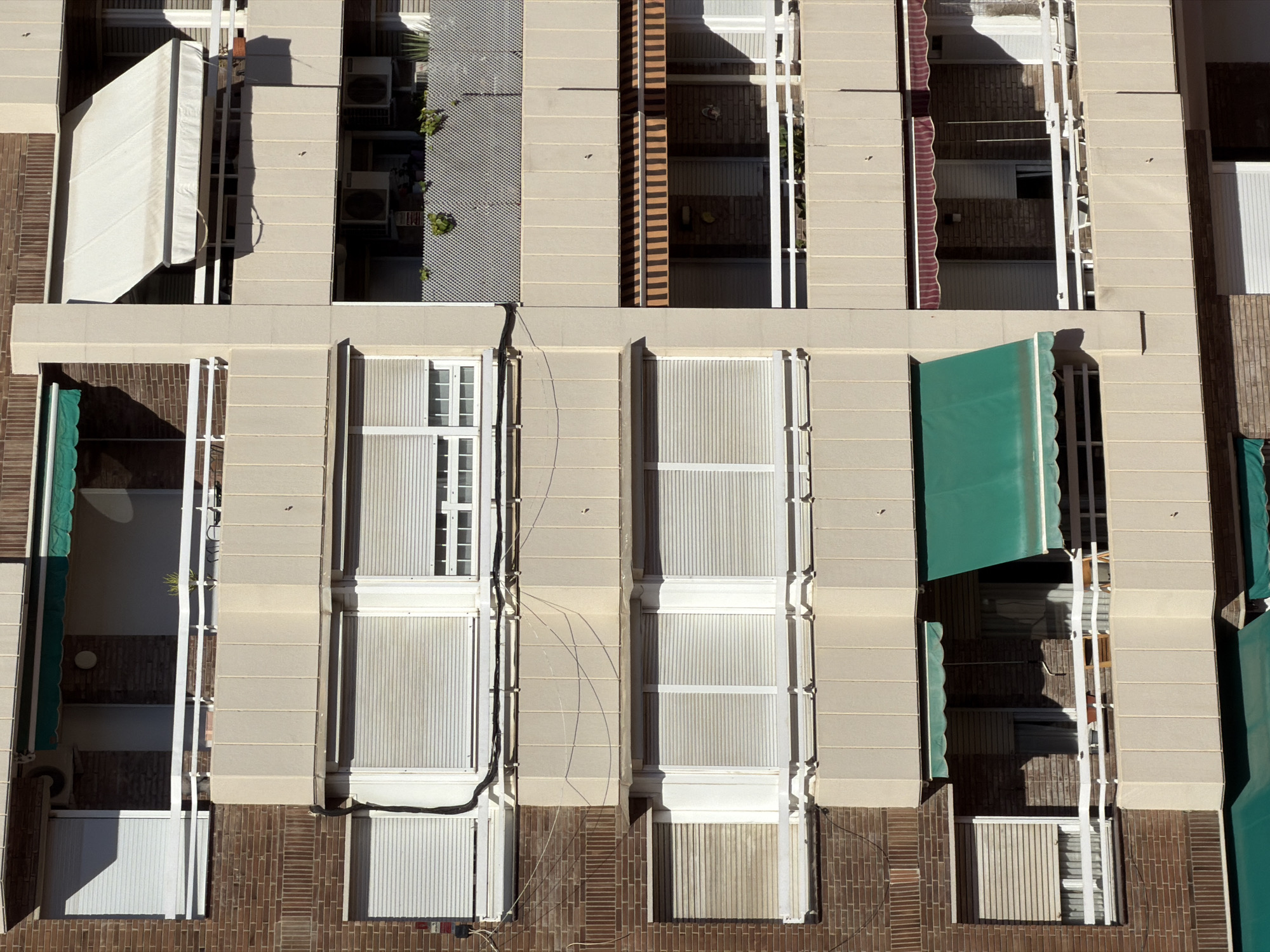 This is a high-angle, exterior shot of a building's facade, likely in Barcelona, Spain, judging by the architectural style. The building features multiple stories with a mix of light-colored masonry and dark brickwork. A prominent feature is the presence of numerous windows, many of which are covered by white or green retractable awnings, some of which are partially deployed, suggesting a sunny day. The awnings are scalloped at the edges.
The windows themselves are mostly covered by white, slatted blinds, creating a uniform look across many of the openings. Some windows appear to be French windows with white frames. In some sections of the building, visible behind the windows or in open balconies, there are glimpses of brick interiors, and the reflection of the sky or other buildings.
There are several air conditioning units mounted on some of the exterior walls, indicating residential or commercial use. There are also some plants visible in small planters or growing on some of the outer walls, adding a touch of greenery. Some electrical conduits and wires are visible running down the facade near some of the windows. The sunlight creates distinct shadows, indicating it is likely daytime, and the sharpness of the shadows suggests it is mid-morning or afternoon. The overall impression is one of urban residential architecture bathed in sunlight. No people are visible in the image. There is no visible text.