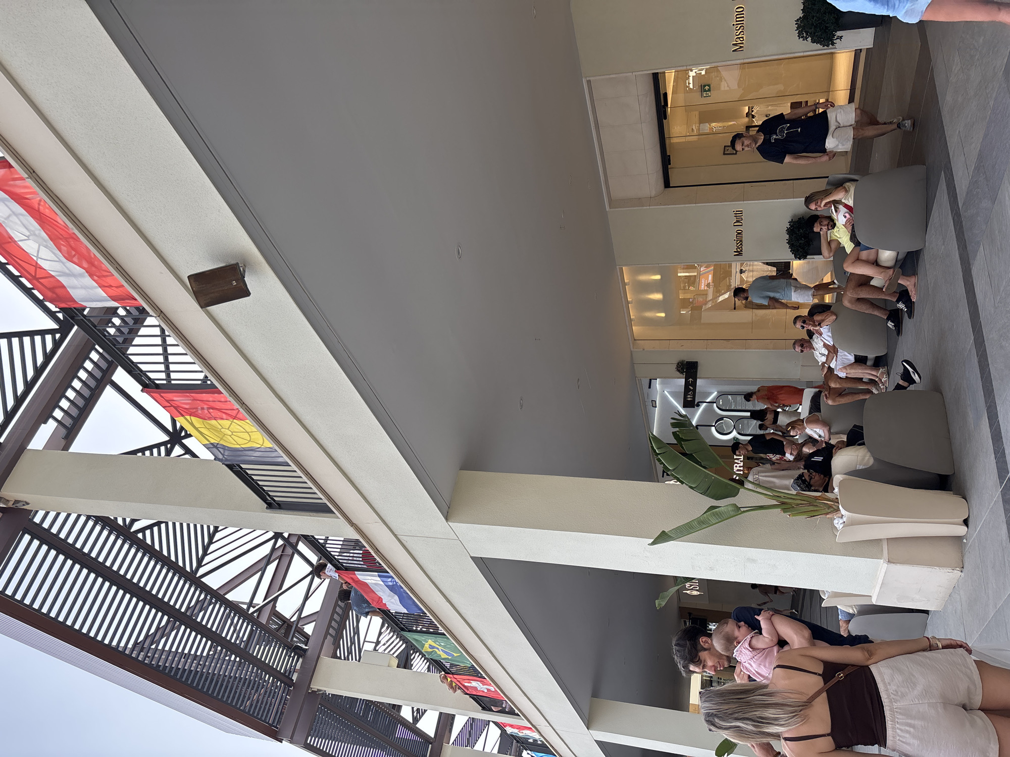 The image shows a street scene with people outside a shopping area. Flags are visible on an upper level, including what appears to be the Austrian flag, the Belgian flag, and the flag of the Netherlands. Several people are sitting in chairs on a lower level, some of them appearing to be in conversation. A man is carrying a baby on the left side of the image. Shops with signs like "Massimo Dutti" are visible. The architecture suggests a modern outdoor shopping mall or pedestrian street. The overall environment is bright and seems to be during the daytime. This scene is located in the city of Orihuela, Spain.