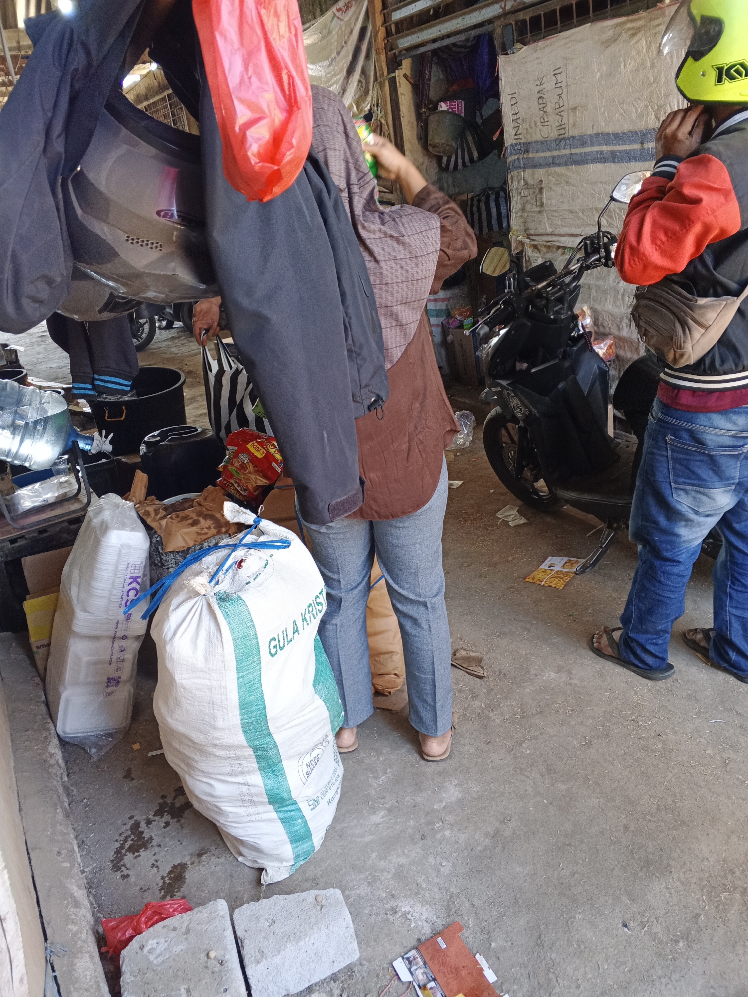 In a bustling outdoor marketplace in Warungkiara, Indonesia, two individuals are preparing for travel. On the right, a person wearing a yellow helmet and a red jacket is adjusting their helmet while seated on a black motorcycle. They are dressed in blue jeans and dark sandals, with a tan fanny pack slung across their chest. To the left, a person in a brown hijab and a brown top is standing near a large white sack of "GULA KRIST" (crystal sugar), with a red plastic bag hanging nearby. Their legs are clad in gray pants and tan sandals. The ground is a mixture of concrete and dirt, littered with debris. Various items are scattered around, including bags of groceries, plastic containers, and discarded materials, indicating a typical scene of commerce and daily life in this Indonesian city. The background reveals a weathered stall structure with various goods stored within.