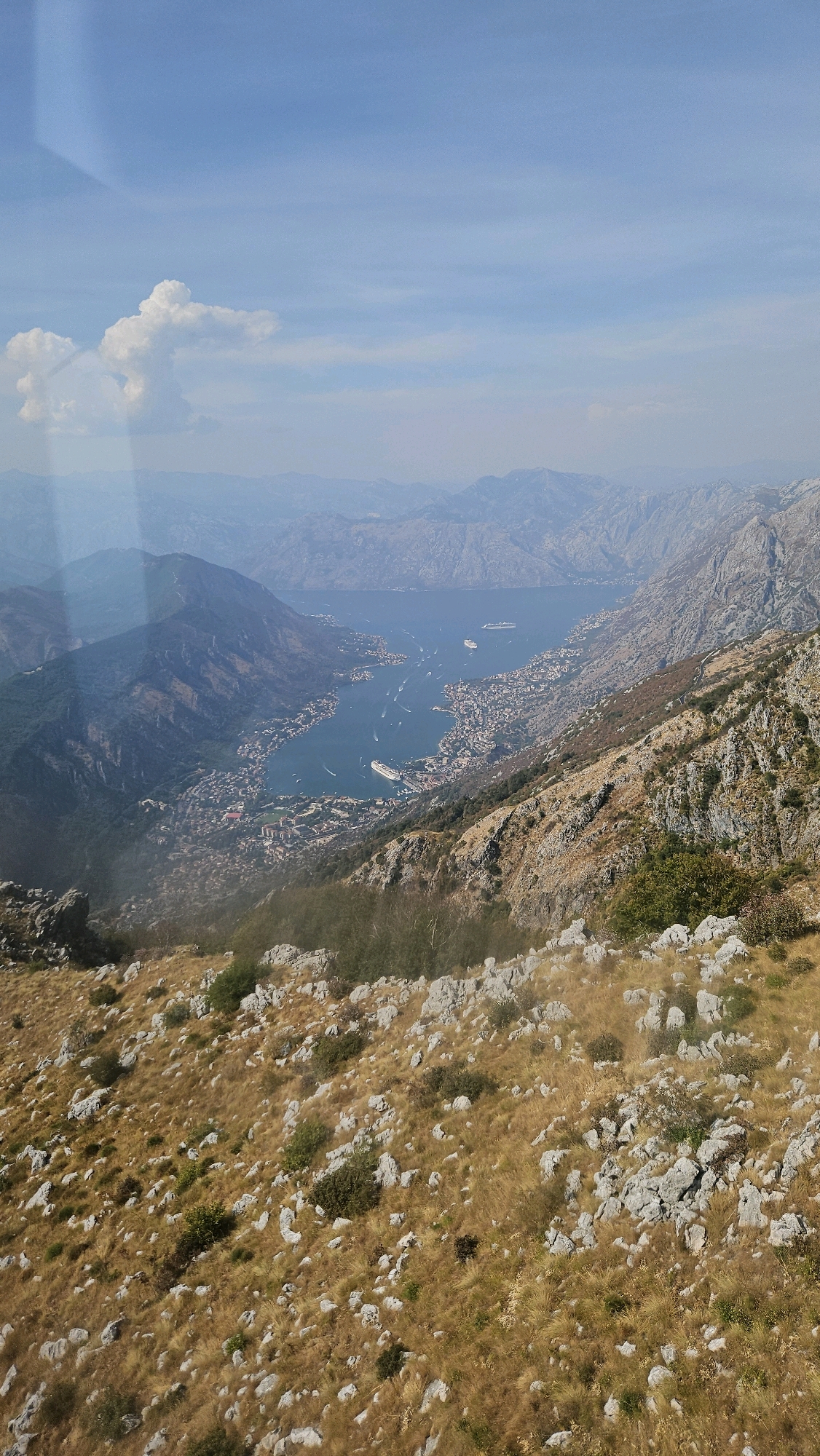 The image is a high-angle aerial view of a bay with a city nestled along its shores, surrounded by dramatic, rocky mountains. The water is a deep blue, dotted with numerous boats of varying sizes, including several large cruise ships. The city appears dense with buildings, concentrated along the coastline and extending up the lower slopes of the mountains. The mountains themselves are rugged and steep, with sparse vegetation on their rocky surfaces, transitioning to more greenery on the lower, gentler slopes. The foreground of the image is a rocky, dry hillside covered in golden-brown grass and scattered rocks, suggesting a Mediterranean or similar arid climate. The sky is a clear blue with a few wispy clouds, and there are visible light flares in the upper left quadrant, likely from a lens or window. The overall impression is a stunning natural landscape integrated with a well-developed coastal city.