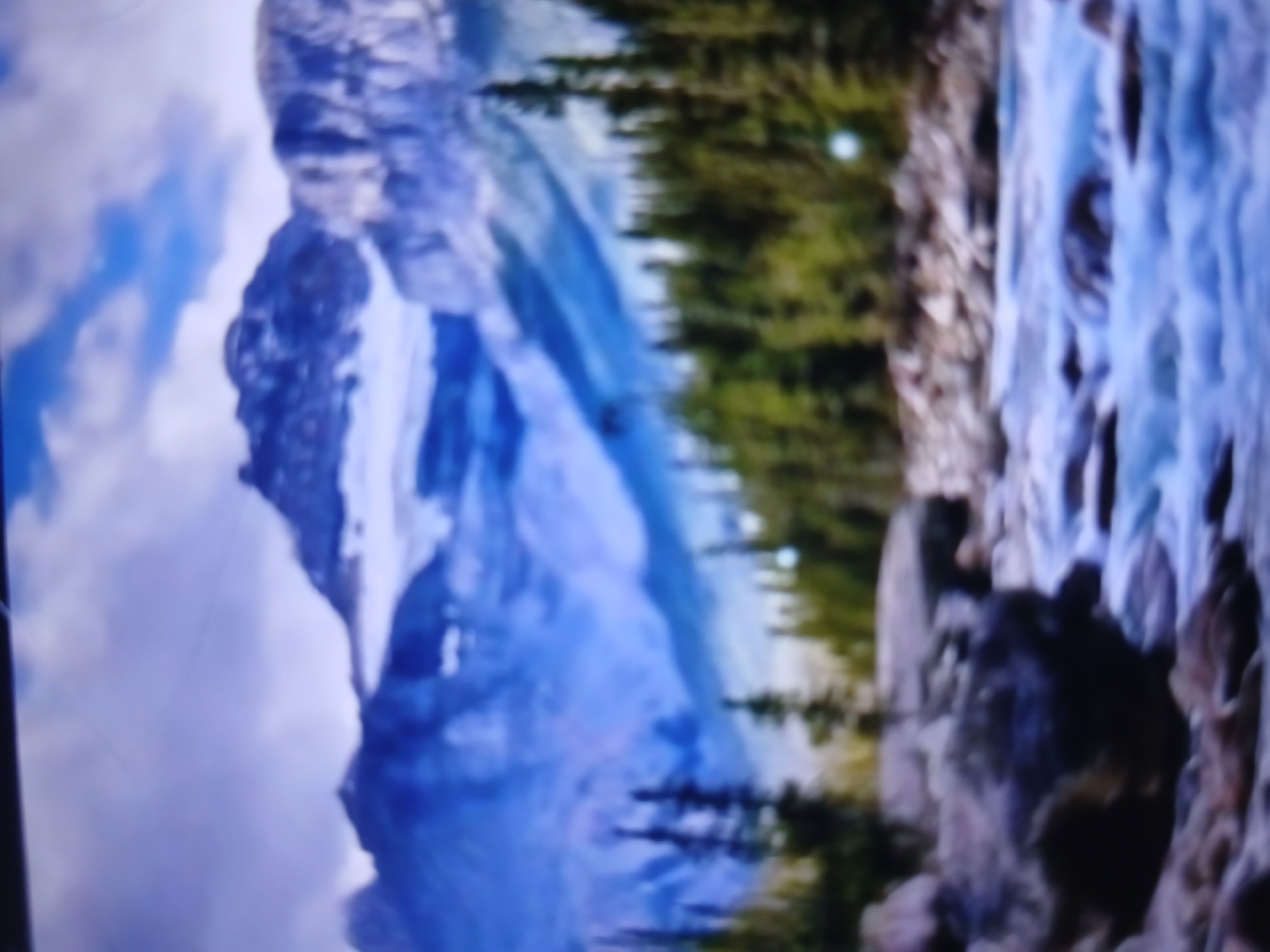 The image displays a blurry, wide-angle view of a mountainous landscape. In the foreground, a rocky riverbed with flowing water is visible. The water is a bright blue, suggesting it is clear and possibly cold. Large, dark rocks are scattered throughout the river and along its banks.
Further back, a dense forest of green coniferous trees lines the banks of the river. The trees appear tall and mature, creating a thick canopy.
The background is dominated by a majestic mountain range. The mountains are snow-capped, with large areas of white snow and ice contrasting with the darker rock faces. The sky above is partly cloudy, with patches of blue visible between the white clouds.
The overall impression is of a wild and natural environment, likely a remote wilderness area. There are no visible signs of human habitation or activity in the immediate scene. The blurriness of the image makes it difficult to discern fine details, but the general composition suggests a scenic natural vista.