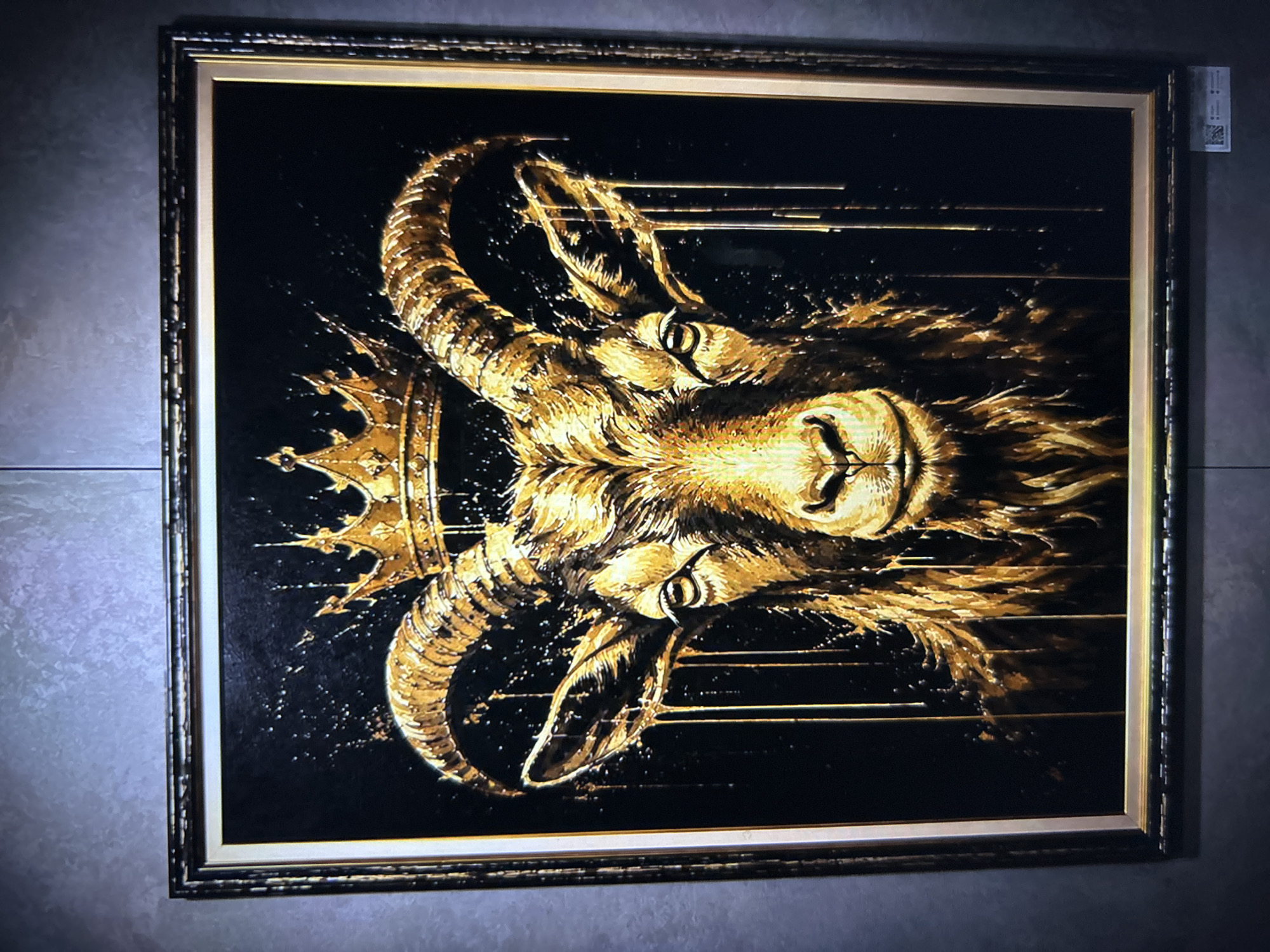 A framed painting of a golden goat wearing a crown is displayed on a textured wall. The goat's head is prominently featured, with its eyes open and looking directly at the viewer. Its horns are large and curved, adorned with golden drips that extend down the sides of its face. The background is black and speckled with gold, giving the impression of a splash effect. The painting is encased in a dark, ornate frame. Below the painting, on the wall, a small white QR code sticker is visible.