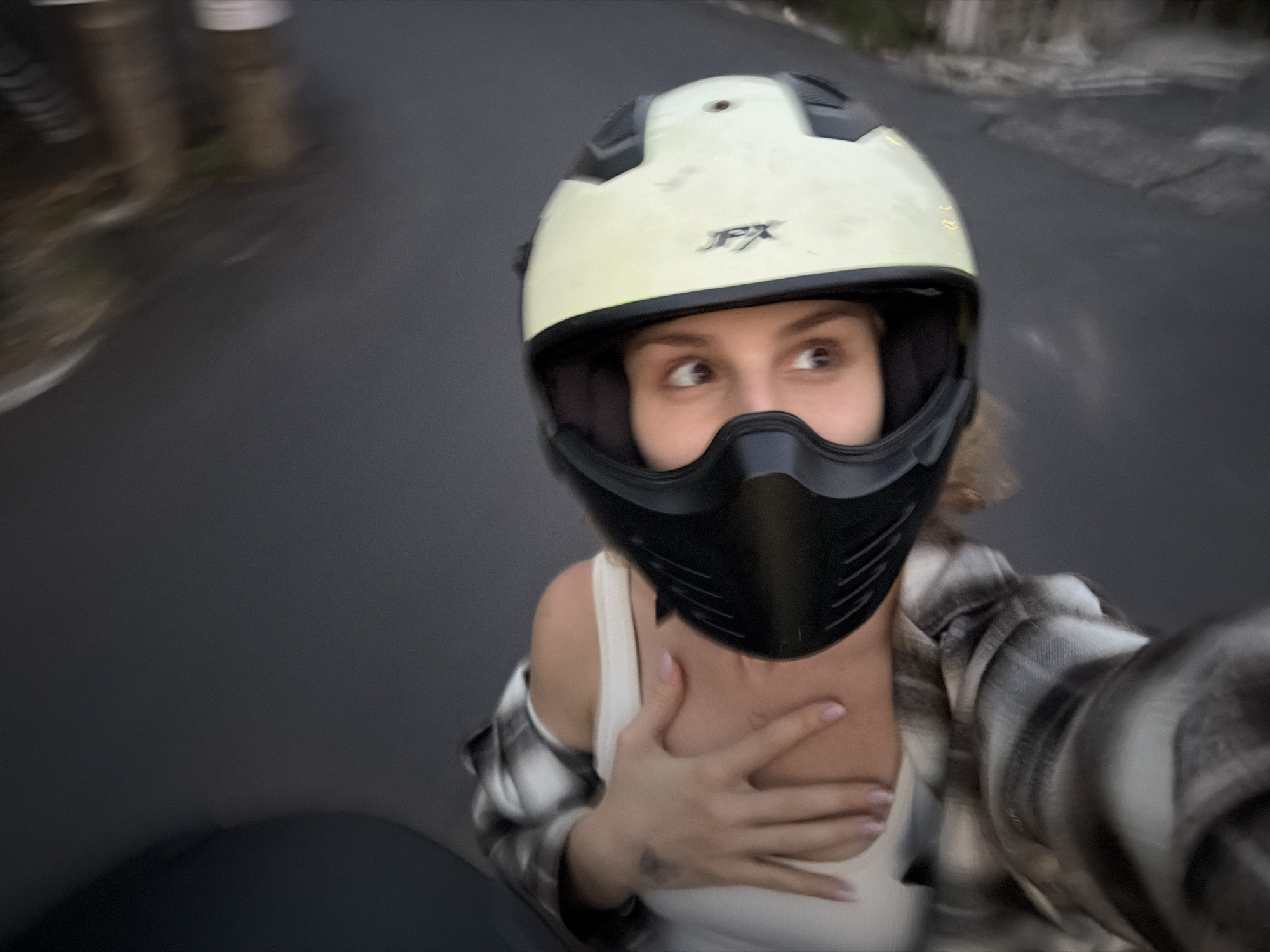 A person wearing a cream-colored JFX motorcycle helmet with a black face mask attachment is riding a motorcycle on an asphalt road. The person is wearing a white tank top and a brown and white plaid flannel shirt. The person's right hand is resting on their chest. The background is blurred, suggesting motion, and includes indistinct vegetation on the side of the road. Based on the information provided, the location is identified as North Kuta, Indonesia.