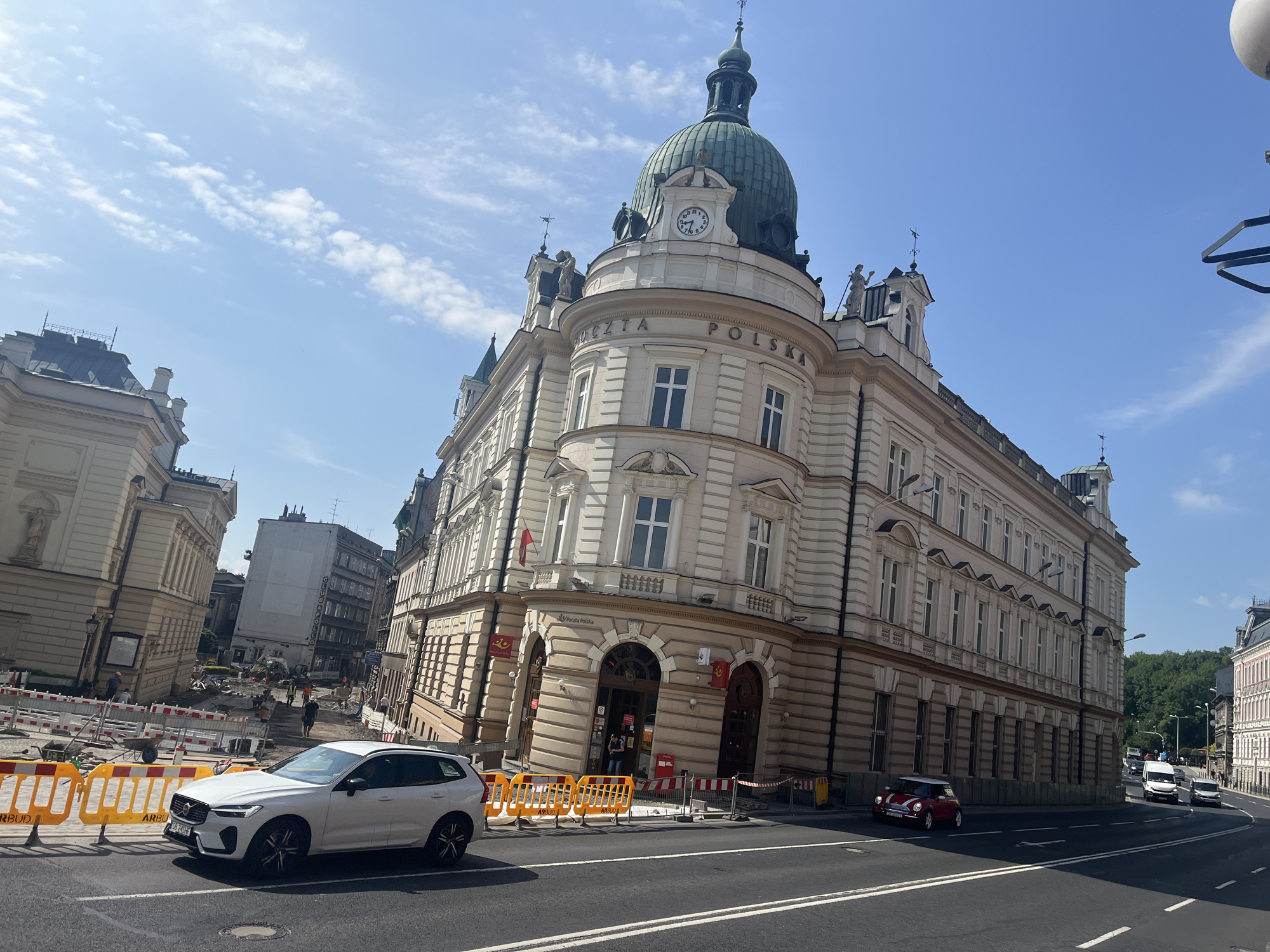 An eye-level, medium shot captures a European city street scene on a clear, sunny day. The focal point is a large, ornate, multi-story building with a green domed roof and a clock tower, identified by the signage "POCZTA POLSKA" (Polish Post). The building's facade is light-colored with numerous windows and decorative architectural elements. In front of the building, construction barriers and equipment indicate ongoing street work.
To the left of the main building, a white Volvo SUV is parked and facing right, partially obscuring some of the construction zone. Further down the street, several other buildings are visible, with a variety of architectural styles. On the right side of the street, a red Mini Cooper is driving away from the viewer. A line of trees can be seen in the background on a hill. The sky is blue with scattered white clouds. Several pedestrians are present on the sidewalks and in the construction area.