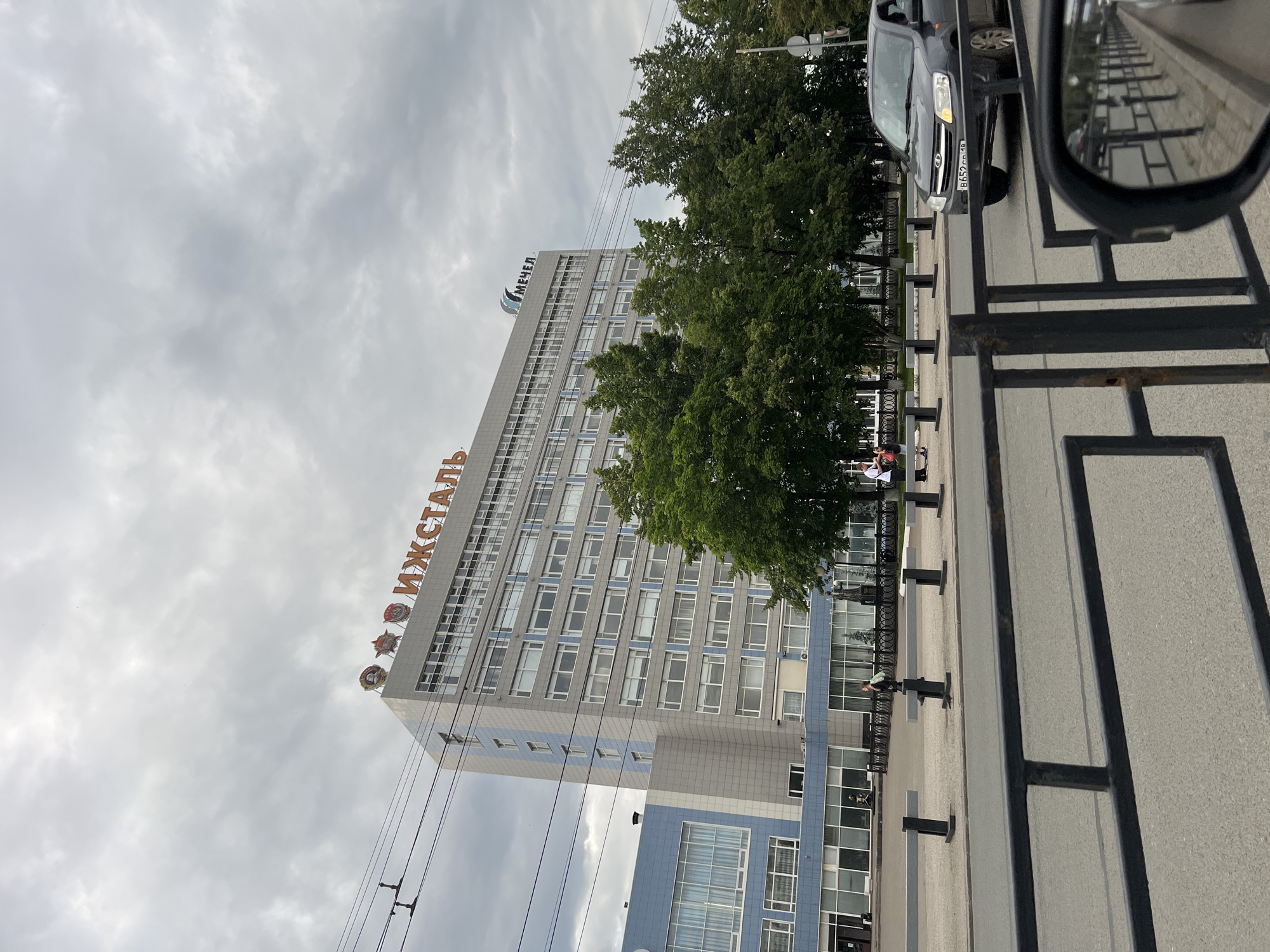 The image shows a view of the IZHMETAL building in Izhevsk, Russia, under a cloudy sky. The building is tall and grey with many windows. On the roof, there are orange letters spelling "ИЖМЕТАЛ" and a logo that says "МЕЧЕЛ." In front of the building, there are trees and a street with a dark-colored car driving on it. There are also a few people walking on the sidewalk. The overall scene depicts a typical urban environment.