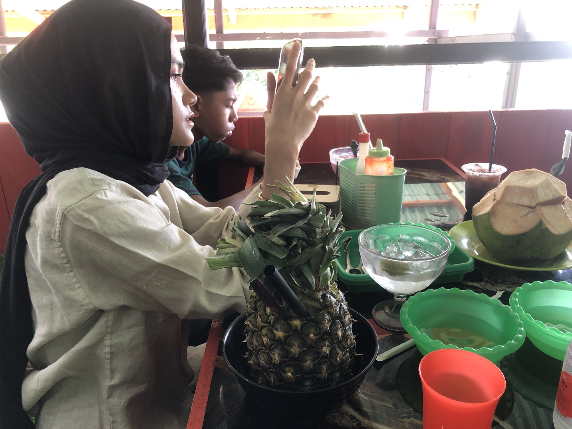 In a brightly lit interior, possibly a restaurant, a person wearing a black hijab and a light-colored, long-sleeved shirt is sitting at a table, holding a cell phone in their right hand. A younger person is seated to their left. On the table, there is a whole pineapple in a black bowl with two straws sticking out, along with multiple green bowls, a clear glass with ice, a drink in a plastic cup with a straw, and a whole coconut. A green plastic container holds bottles and condiments. The location is identified as the city of Lancok, Indonesia.