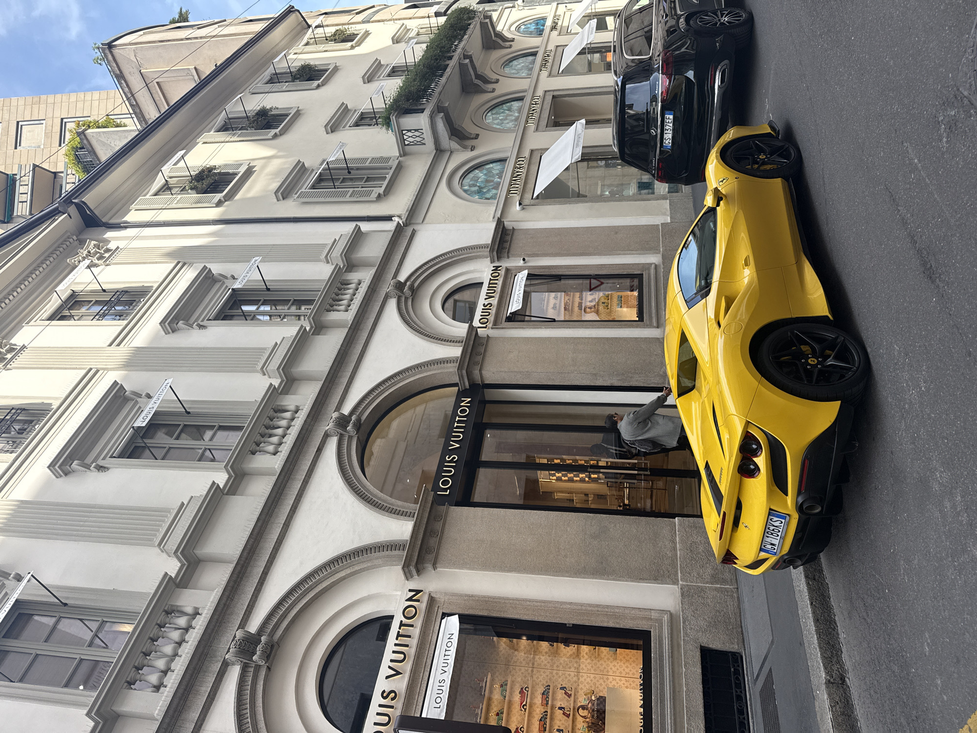 The image shows a street in Milan, Italy. A yellow Ferrari is parked next to a Louis Vuitton store. In front of the Louis Vuitton store is also a black Mercedes SUV. The building is ornate, with arched windows and signs for Louis Vuitton and Tiffany & Co. hanging above the windows. A man is seen leaning against the Louis Vuitton window.