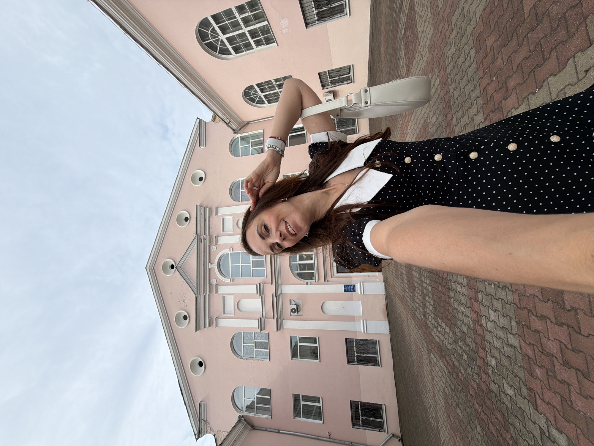 The image is a first-person, wide-angle selfie taken outdoors. A woman with long brown hair is positioned in the foreground, smiling and looking at the camera. She is wearing a black and white polka dot dress with a white collar and gold buttons, and carrying a light-colored handbag over her left shoulder. Her right arm is extended towards the camera. Behind her, a large, pale pink building with arched windows and ornate architectural details dominates the background. The building appears to be several stories high. The ground in front of the building is paved with reddish-brown and gray interlocking bricks. The sky above is overcast with some visible clouds. The scene appears to be a public space, possibly in front of an educational institution or administrative building. The location is identified as Birobidzhan, Russia.