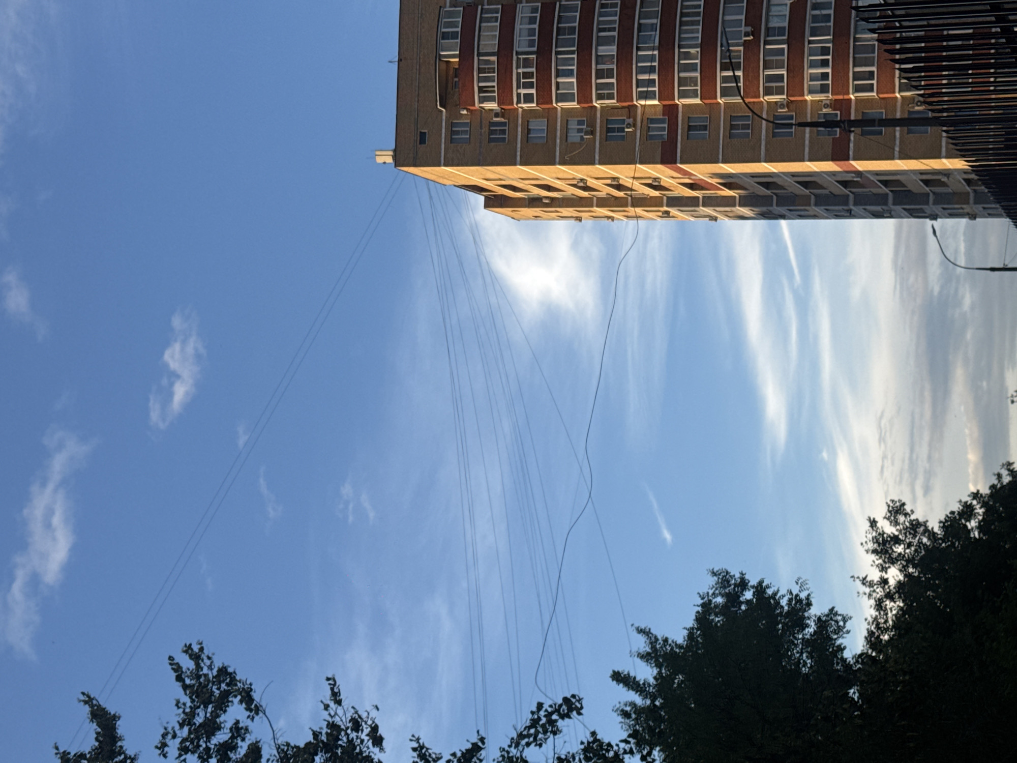 This is an exterior, low-angle shot of a modern, multi-story apartment building constructed with light brown and red brick. The building features numerous windows with white frames and some with balconies that have red detailing. The sky is a vibrant blue with scattered white clouds, suggesting it is daytime. Several power lines are visible crisscrossing the sky in the foreground and middle ground. On the left side of the image, dark green foliage from trees frames the scene. In the lower right corner, a black metal fence is visible, along with streetlights. The building's facade is illuminated by sunlight, casting a warm glow. The overall impression is of an urban residential area under a clear sky.