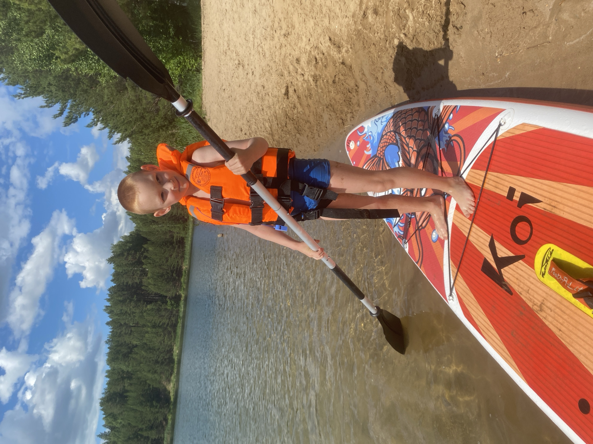 In this vertical, outdoor shot taken in Novyi, Russia, a young boy with fair skin and light hair is the central subject. He's wearing an orange life vest over blue shorts and is standing on a paddleboard. His left hand grips a black paddle, and he's looking at the camera with a slight smile, conveying a sense of readiness or mild amusement.
The paddleboard, predominantly red with orange stripes and a blue and orange fish design, rests on the shallow edge of a body of water. The water appears murky and calm, reflecting the light. Behind the boy, a sandy beach transitions into a dense forest of green trees, indicative of a natural, possibly recreational, setting. The sky above is a vibrant blue, dotted with fluffy white clouds, suggesting a pleasant, sunny day, likely in the afternoon given the lighting.
The overall atmosphere is relaxed and summery, hinting at a day of outdoor activity. The boy's posture and the paddle suggest he's either about to start paddling or has just arrived at the shore. The image captures a moment of childhood adventure by the water. There's a small yellow object with some text on the paddleboard, but the writing is too blurry to decipher. The word "KO" is prominently visible on the paddleboard in black lettering.