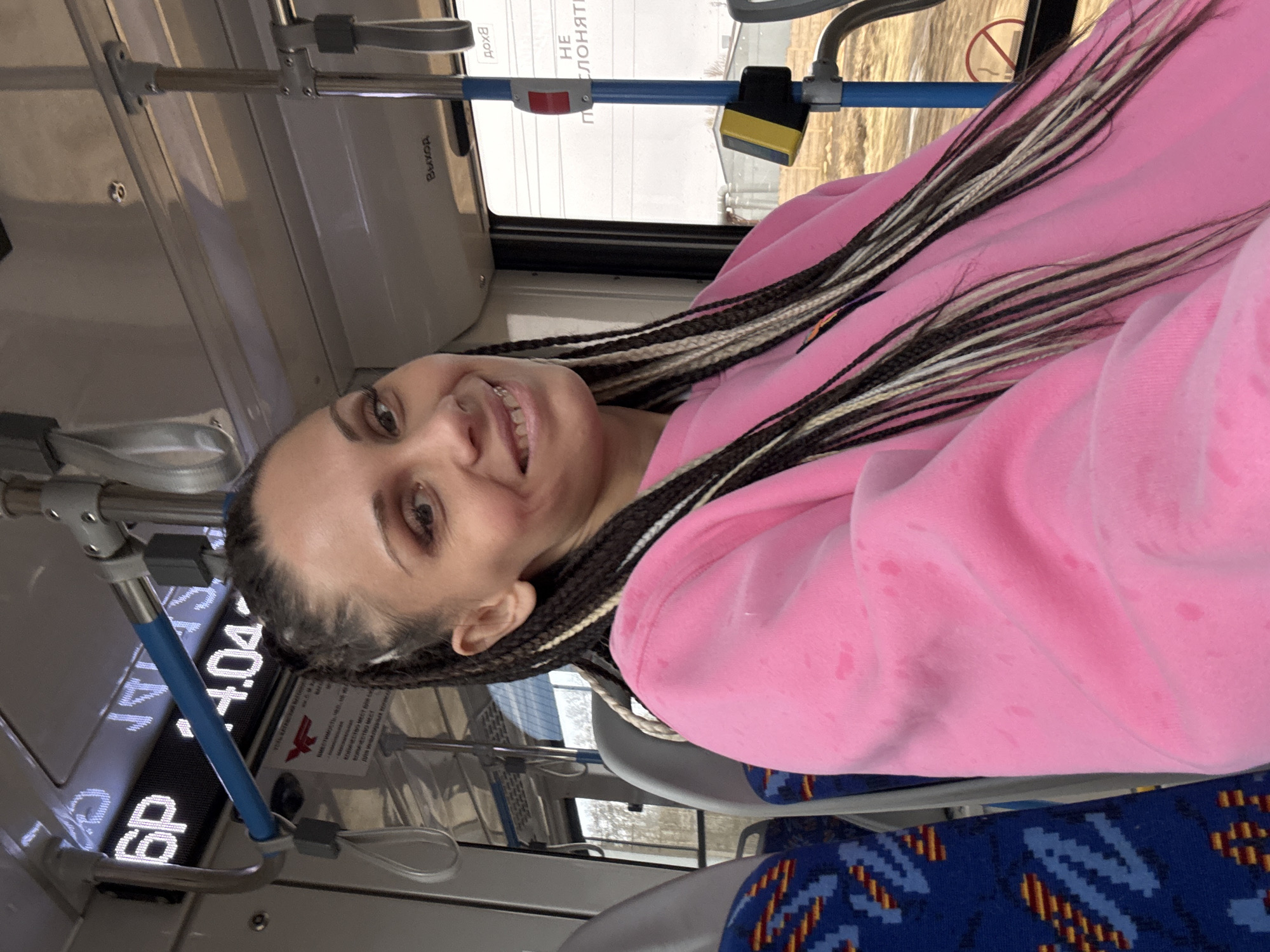 A selfie of a woman on a bus in an unknown city. The woman is wearing a pink sweater and has long black and blonde braids. She is sitting in a seat with a blue and yellow pattern. The bus interior is visible, including metal handrails, windows with Russian text, and an electronic display showing "6P 4.04". The scene appears to be inside a public transportation vehicle during daytime.