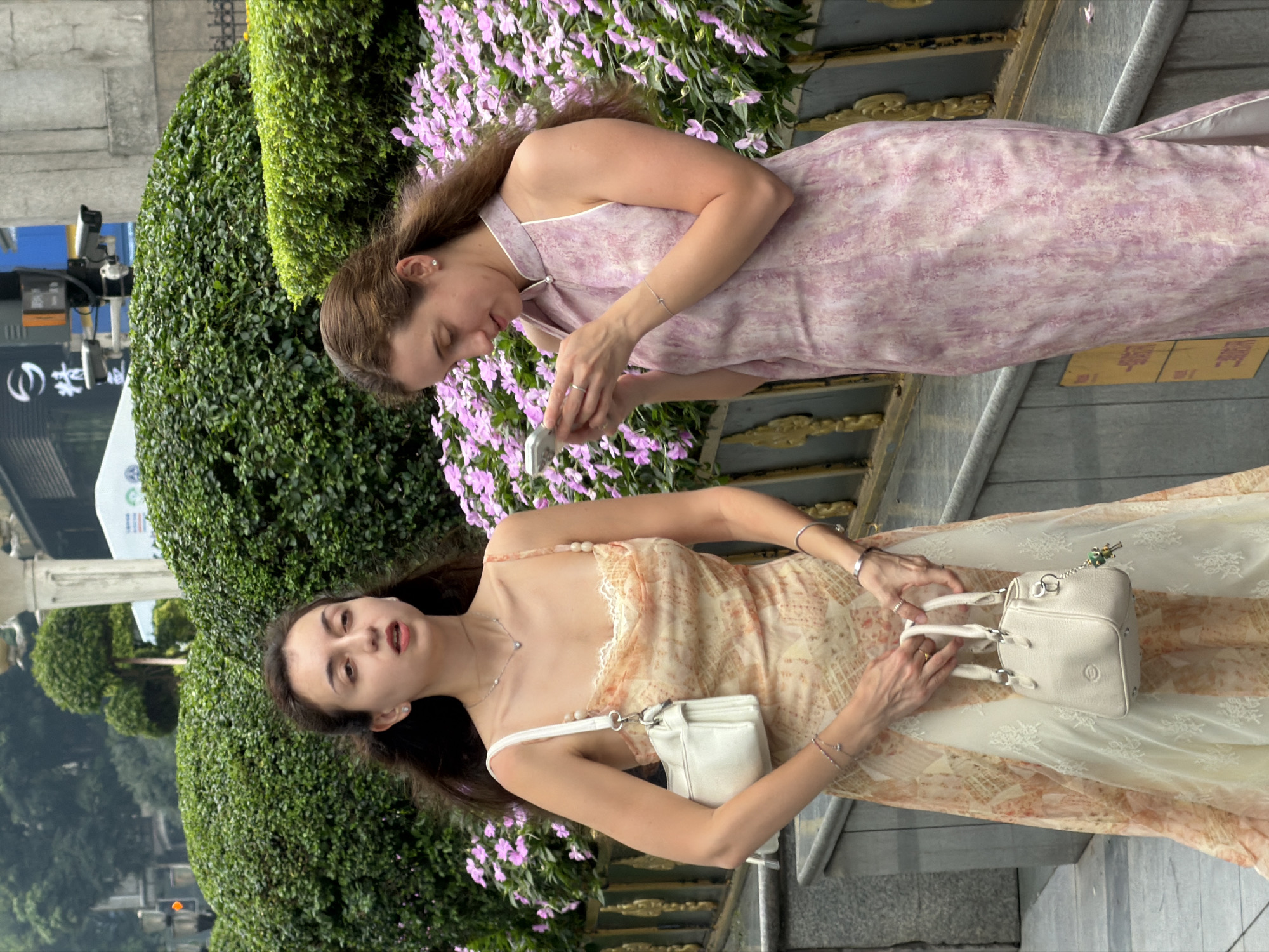 In Yuzhong District, China, two young women stand near a lush green hedge and a bed of pink flowers. The woman on the left, with dark hair, wears a light-colored, patterned dress with spaghetti straps and carries a white handbag. She is looking forward with her mouth slightly open. The woman on the right, with brown hair, is dressed in a sleeveless pink dress with a high collar. She is looking down at her smartphone, which she is holding in both hands. She is wearing a ring on her left hand and a delicate bracelet on her right wrist. Both women are adorned with necklaces and earrings. The background shows a blurred cityscape with architectural elements and traffic signals, suggesting a public urban setting.