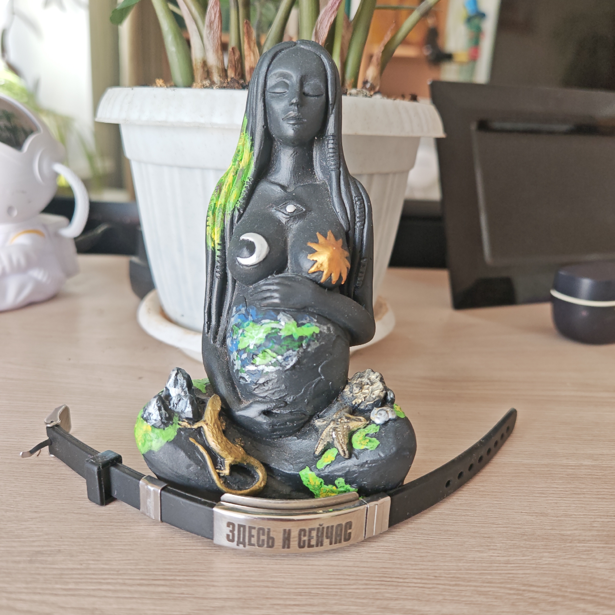 On a light brown wooden surface is a statue of a seated, pregnant female figure, painted dark gray. The figure's abdomen is painted as a globe, and she has a crescent moon on her left breast and a sun on her right breast. The figure's hair is partially painted green. At the base of the statue, near the feet, are painted miniature reptiles and a starfish. In front of the statue is a black wristwatch band with a silver plate that reads "ЗДЕСЬ И СЕЙЧАС" in Russian, which translates to "HERE AND NOW". In the background, behind the statue, is a white potted plant, a white astronaut figurine, and a black square object. The image location is Rostov, Russia.
