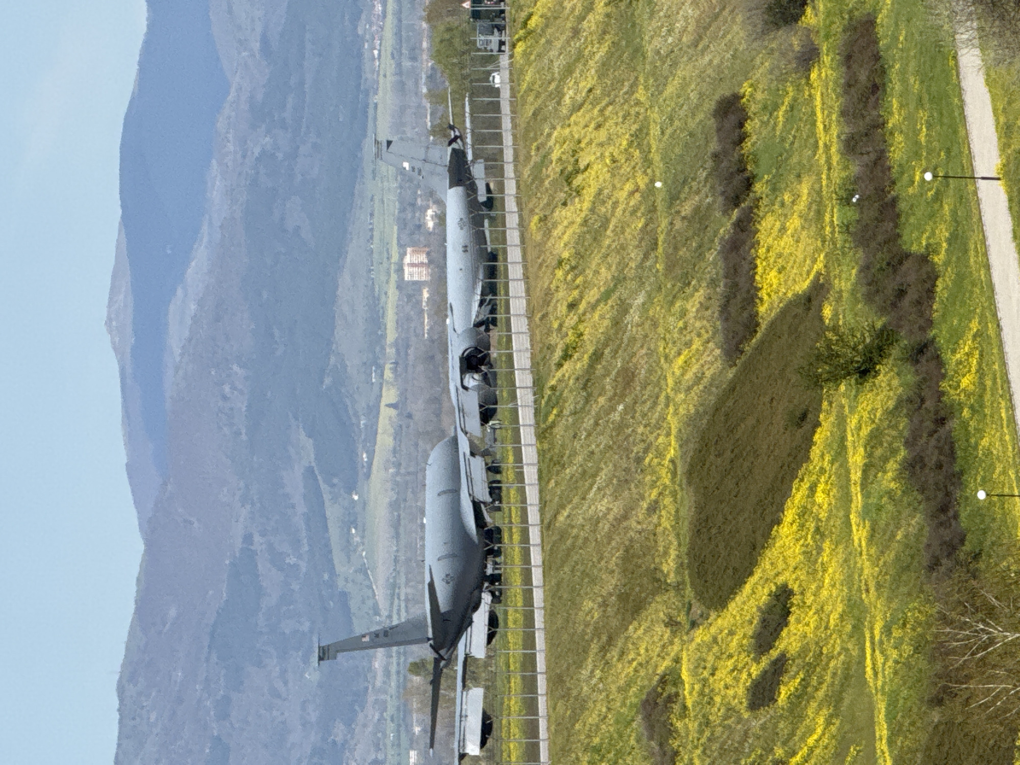 The image, oriented horizontally, showcases two large, gray military transport aircraft parked on an airfield, likely an apron or a taxiway. The aircraft are positioned one behind the other, facing left. Their design, characterized by a high-wing configuration and four engine nacelles (typical of turboprop engines), suggests they are C-130 Hercules or similar cargo planes.
In the foreground, to the right of the aircraft, there is a vibrant expanse of green and yellow vegetation, indicative of a field covered in flowering plants, possibly rapeseed or mustard, suggesting springtime. A narrow, light-colored path or road runs along the right side of this field, featuring several modern streetlights. A metal fence, possibly a chain-link barrier, separates the aircraft parking area from the adjacent field.
The background is dominated by a majestic range of blue-tinged mountains, receding into the distance under a clear, bright blue sky. Some scattered buildings and signs of urban development are visible in the valley between the aircraft and the mountain range.
The scene takes place during daytime, under clear weather conditions, with soft shadows suggesting either late morning or late afternoon. The aircraft appear stationary, and no people are visible in the frame. Visible markings on the aircraft, including a partial flag design on the tail of the rear aircraft and other insignia on the fuselage of the front aircraft, are present but not clearly legible at this resolution. The context provided indicates this location is Sofia, Bulgaria, suggesting an airport or airbase in the region.