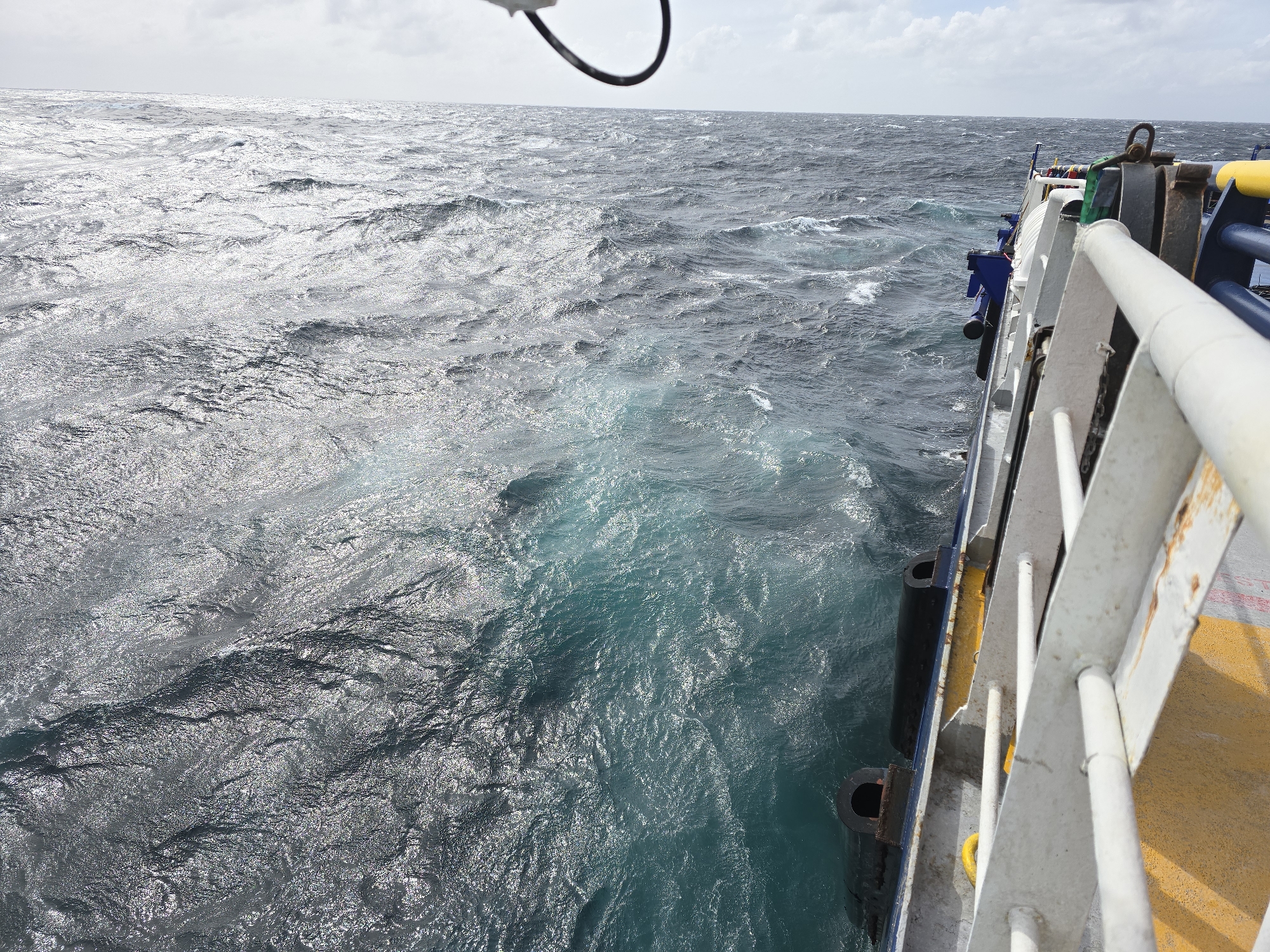 The image depicts a view from the deck of a ship at sea. The ocean water is choppy and displays varying shades of blue and gray, reflecting the sky. The ship's railing and parts of its structure are visible on the right side of the frame. The location is the open ocean; the city and specific location are unknown. There are no people visible. The scene primarily shows the interaction between the ship and the ocean environment.