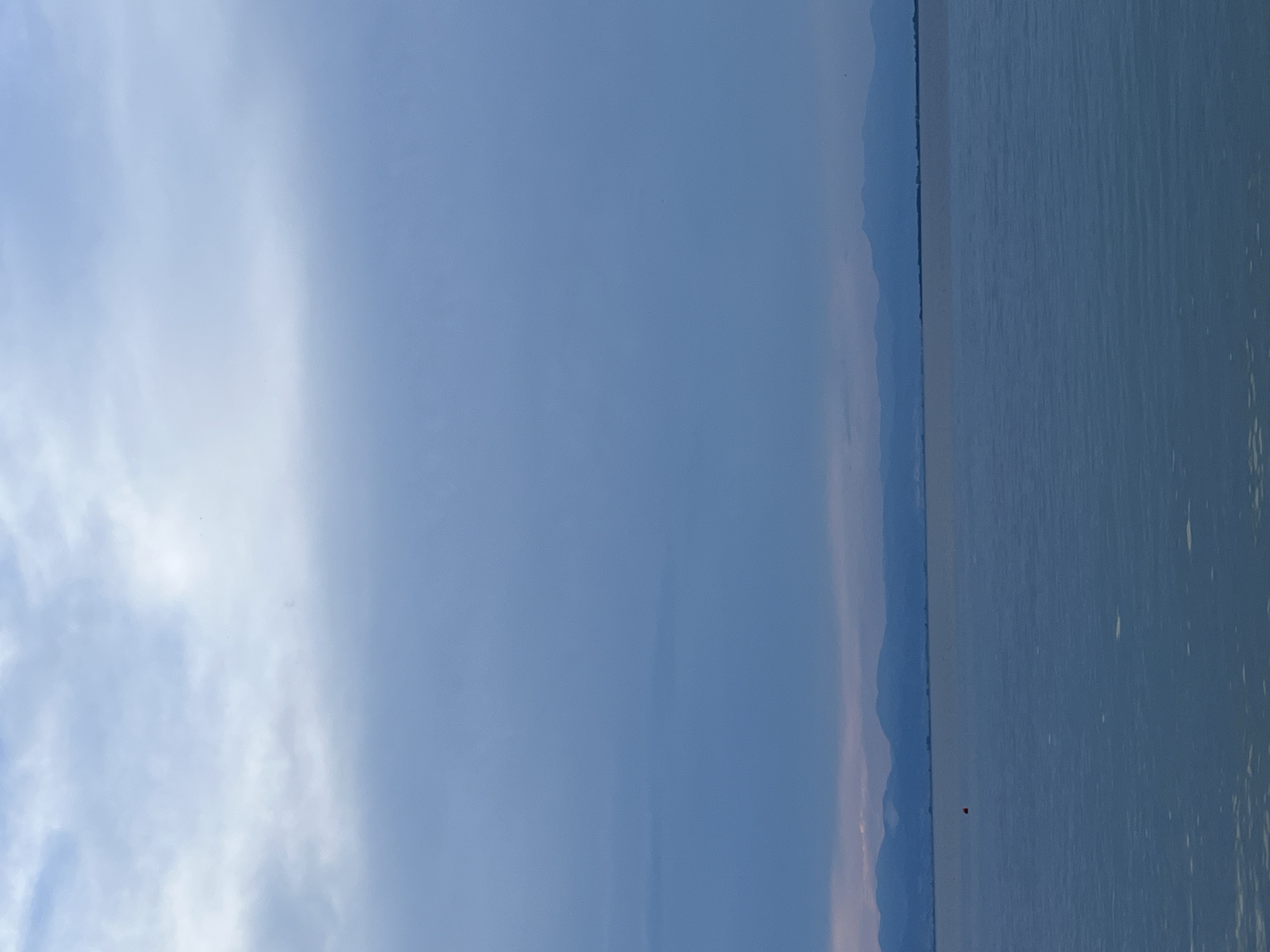 A serene, wide-angle view captures a calm body of water, likely the sea or a large lake, stretching towards a distant horizon where a range of blue mountains meets a soft, overcast sky. The water reflects the muted tones of the sky, creating a tranquil and expansive atmosphere. In the middle distance, a small, bright orange buoy floats on the water's surface, adding a single point of vibrant color against the predominantly blue and gray palette. The overall impression is one of peace and natural beauty, with the mountains providing a majestic backdrop to the still water. This scene is consistent with the coastal landscapes found around Thassos, Greece, known for its picturesque natural settings and clear waters.