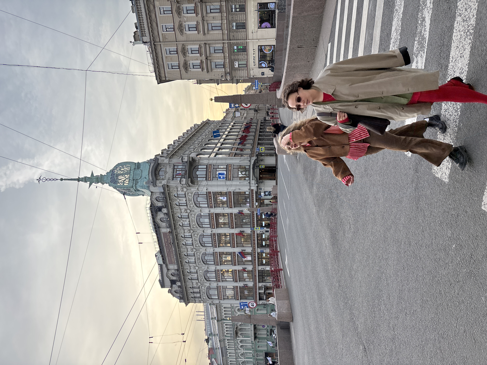 Two women stand on a crosswalk in front of a prominent building in Saint Petersburg, Russia. The building is a large, ornate structure with a green-domed tower, identified as the Singer House (Dom Knigi). The woman in the foreground wears a brown jacket and pants, and the woman behind her wears a tan coat. Both are wearing sunglasses. The background includes a portion of Nevsky Prospect, the city's main avenue.