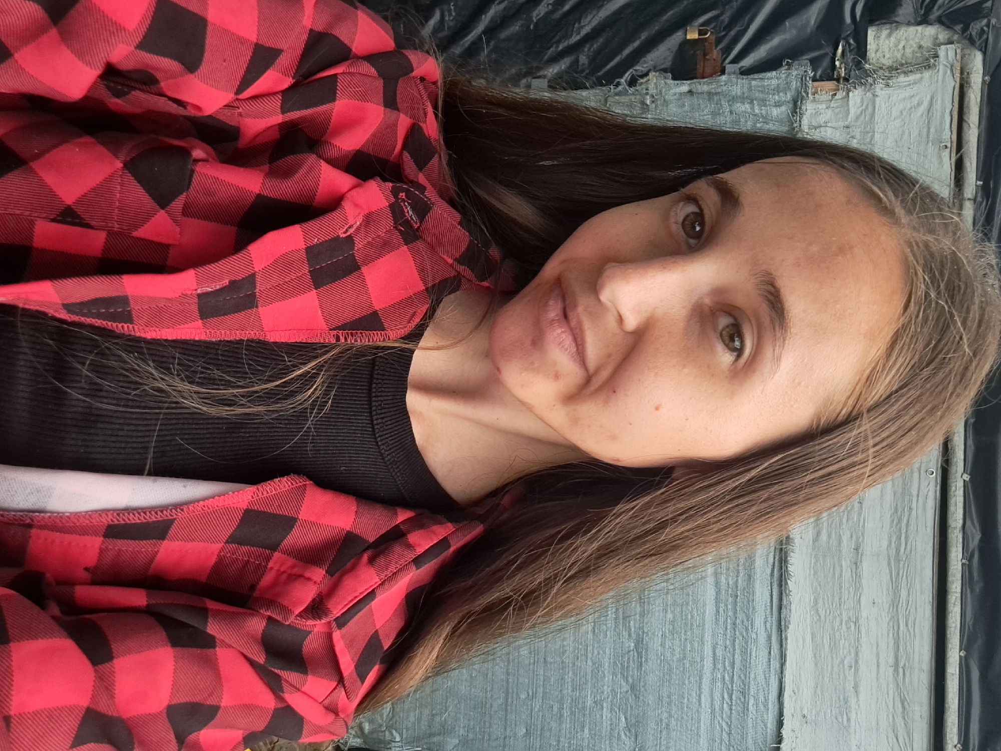 The image shows a close-up, selfie-style photograph of a young woman with long, brown hair. She is wearing a red and black plaid shirt over a black ribbed top. Her expression is neutral with a slight smile. The background appears to be outdoors, with a textured, light blue tarp or siding visible behind her. To the left of the woman, a black tarp is draped over some structure, and a small, brass-colored padlock is attached to a metallic element. The image's context suggests it was taken in Vypasne, Ukraine, though no specific landmarks or environmental details within the city are discernible from the provided view.