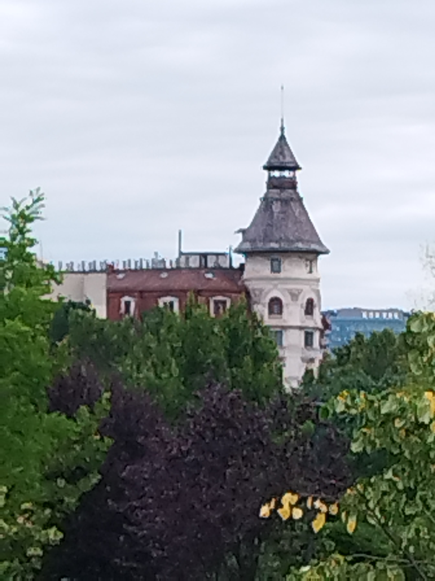 The image shows a view of the Bucharest Stock Exchange building, also known as the Palace of the Romanian Chamber of Commerce and Industry. The building features a distinctive turreted tower and is partially obscured by trees and foliage. The sky is overcast, suggesting a cloudy day. The style of architecture indicates a historical building, likely from the late 19th or early 20th century.