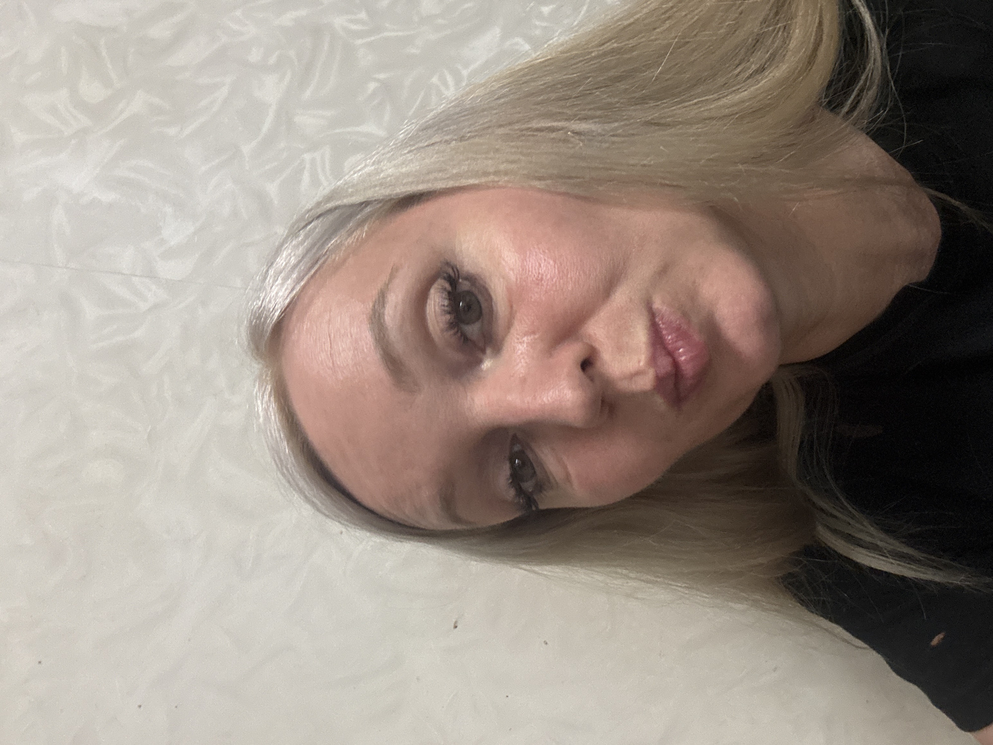 The image shows a close-up, selfie-style photograph of a woman with blonde hair and fair skin. She is wearing a black top and has on makeup, including mascara and lipstick. Her lips are pursed. The background appears to be a textured, light-colored wall. The photograph was taken in the city of Komsomolsk-on-Amur, Russia.
