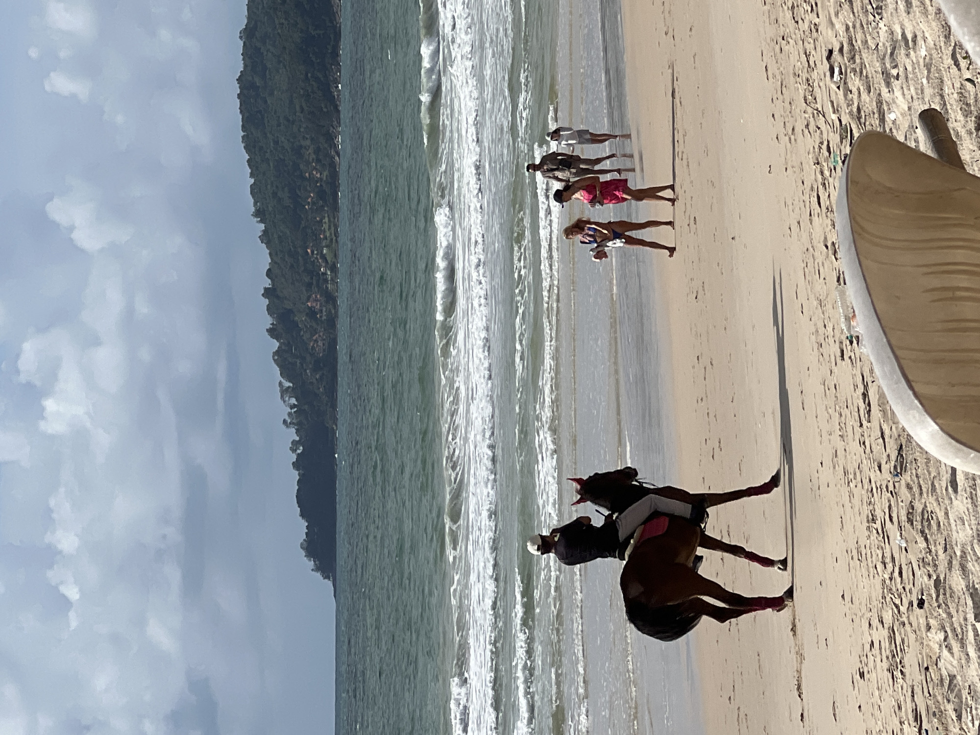 A horse with a rider is walking on a sandy beach near the water. The rider, wearing a black shirt, light pants, and a white cap, is facing away from the camera. The horse is brown with red leg wraps. In the background, several people are walking along the water's edge. One woman is wearing a blue bikini, and another is wearing a pink dress. Further back, two more individuals are visible. The ocean has moderate waves with white foam. In the distance, a lush green, tree-covered hill with some buildings is visible. The sky is cloudy with patches of blue. The sandy beach appears to have some litter. This scene is consistent with a beach in Choeng Thale, Thailand, known for its coastal attractions.