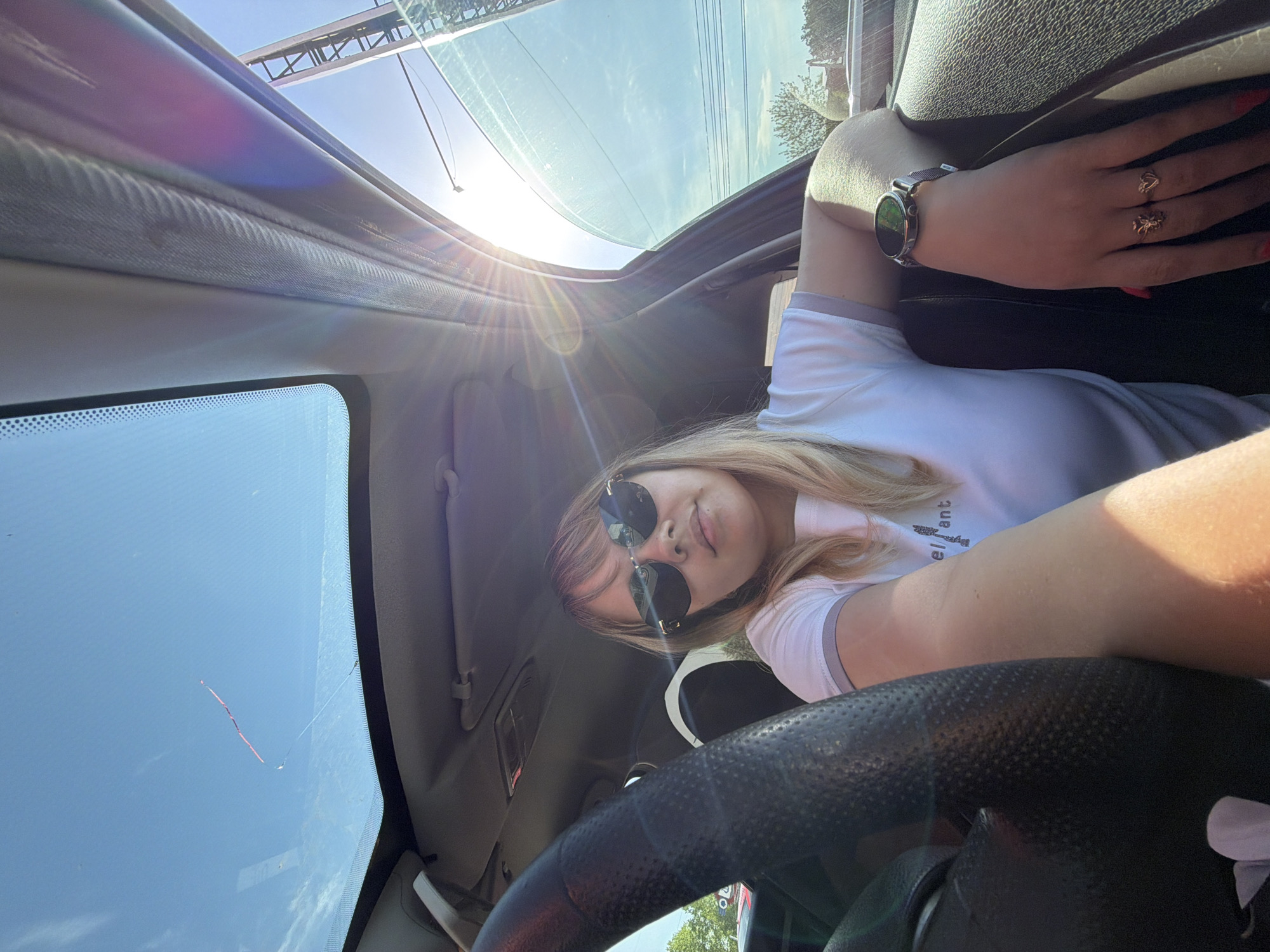 A young woman sits in the driver's seat of a car. She is wearing sunglasses, a white shirt with grey trim, a watch on her left wrist, and rings on her fingers. The car has a sunroof, which is open. The steering wheel is black. The location is Chaikovsky, Russia. The lighting indicates it is daytime.