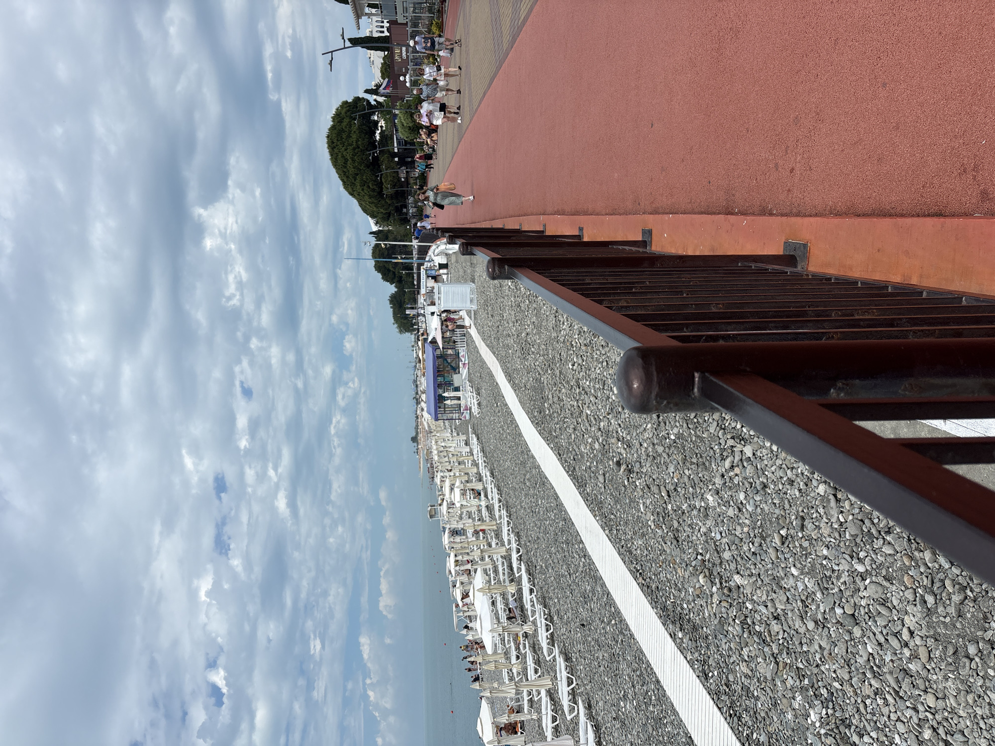 The image depicts a paved walkway running alongside a pebble beach in Sochi, Russia. The walkway is a vibrant red and is bordered by a dark railing in the foreground. On the left side of the walkway, rows of empty white lounge chairs and umbrellas are neatly arranged on the grey pebble beach, indicating a quiet, perhaps early or late, period of activity. The calm sea stretches out to the horizon under a sky filled with dramatic, cloudy formations. A few people are visible walking on the red walkway, with one woman in a green dress carrying shopping bags. Further in the distance, more people are seen on the beach and along the promenade, with buildings and lush green trees forming a backdrop. Russian flags are visible on a building in the distance. The overall impression is of a well-maintained coastal area, likely a popular spot for leisure and recreation.